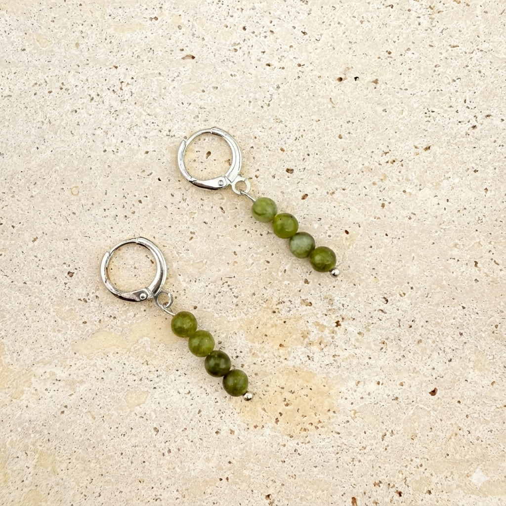 A pair of silver Aking Sinta huggie earrings on a white textured rock; each earring features a vertical strand of four round, dark green Canadian Jade beads suspended from a closed, polished silver hinged hoop.