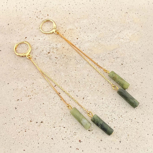 A pair of Aking Sinta gold huggie earrings featuring a dramatic double drop design. Each earring has two delicate gold chains of varying lengths, each ending in a polished Canadian jade column pendant. Displayed on a light-colored natural stone surface