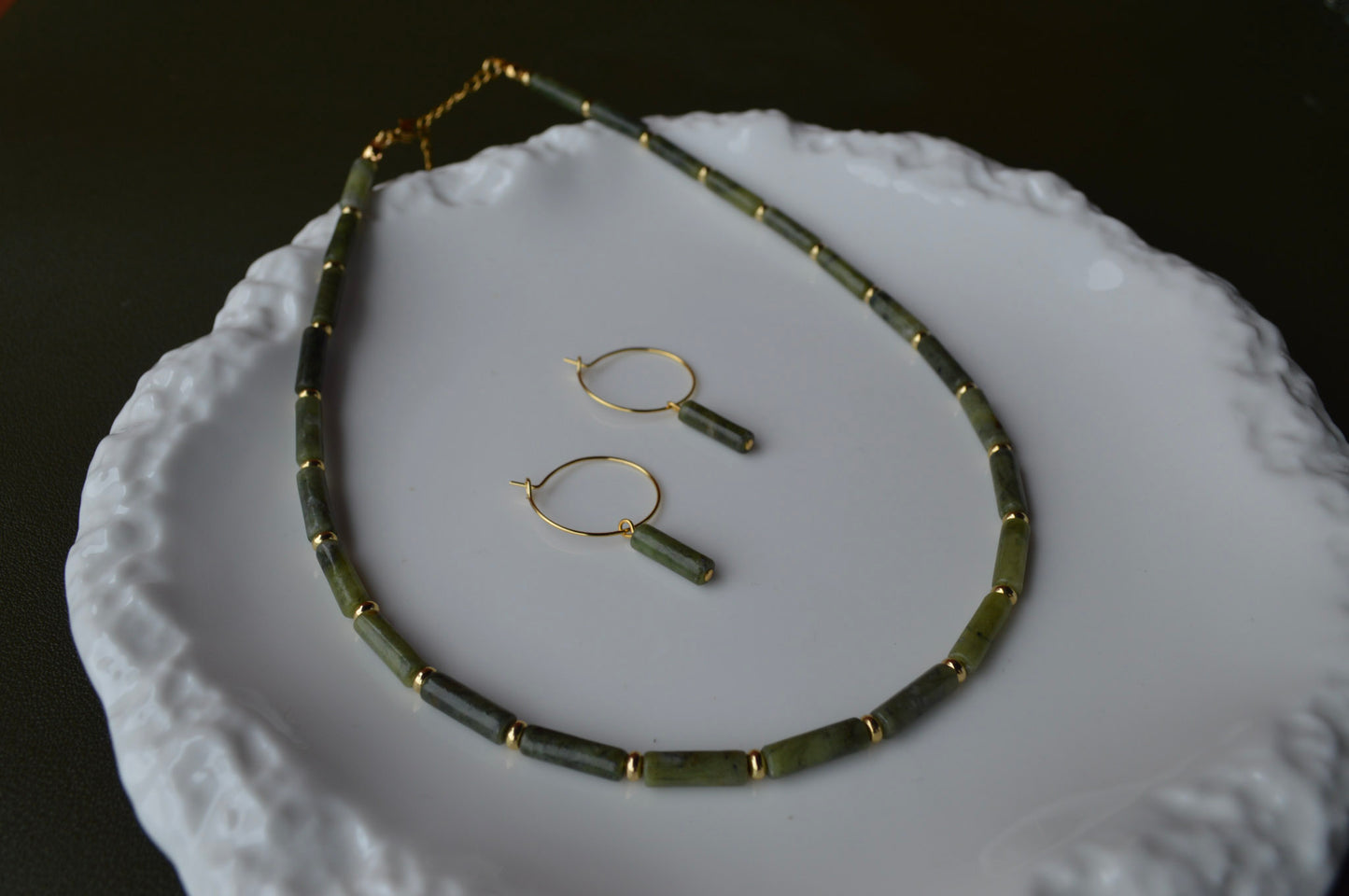 A pair of gold hoop earrings with vertical Canadian Jade column charms resting inside a shimmering Mother of Pearl shell container; the dark green translucent stones contrast beautifully with the iridescent pink and white background.