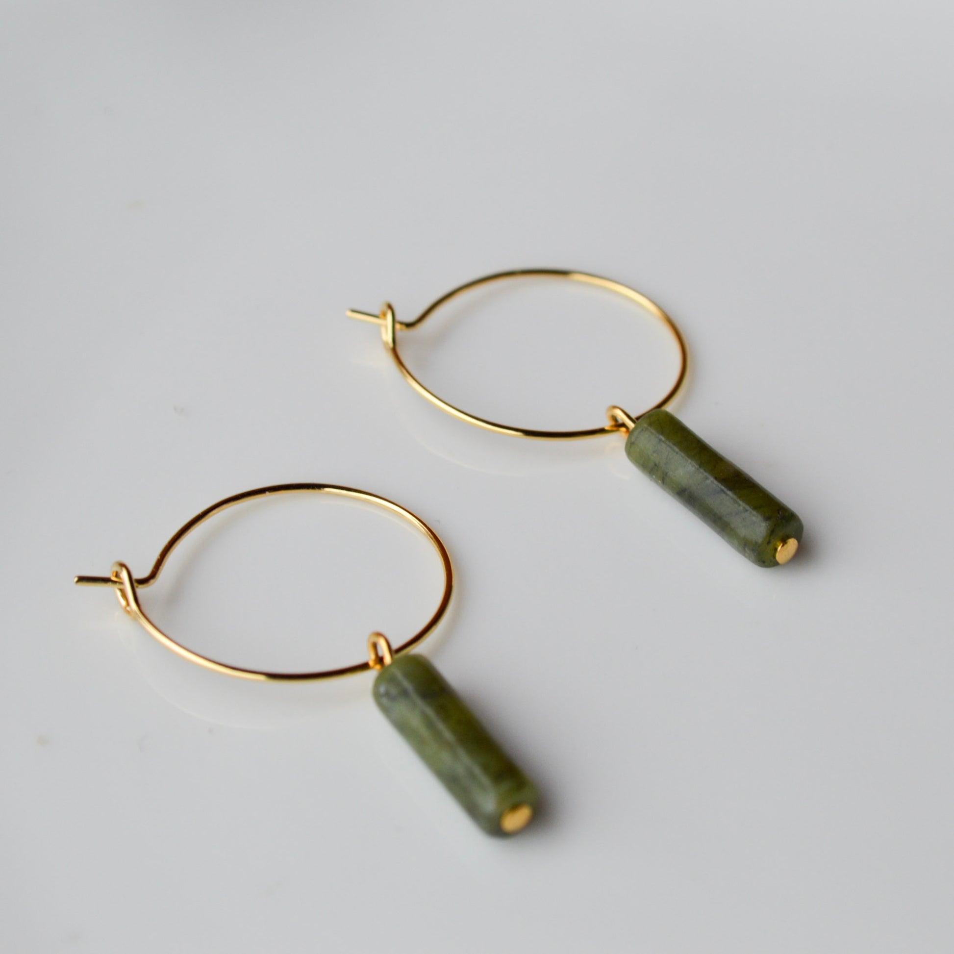 A pair of gold hoop earrings with vertical Canadian Jade column charms resting inside a shimmering Mother of Pearl shell container; the dark green translucent stones contrast beautifully with the iridescent pink and white background.