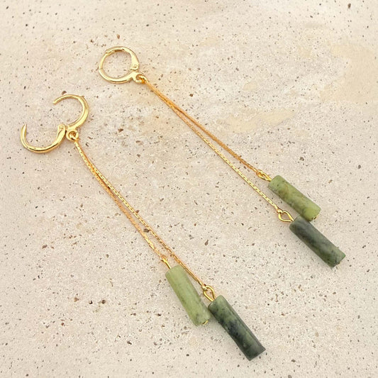 A pair of Aking Sinta gold huggie earrings featuring a Canadian jade double drop design. One earring is open to show the clasp, highlighting two gold chains of different lengths with polished green jade column pendants on a light stone background