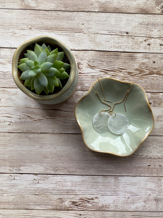 A pair of Aking Sinta gold ball chain threader earrings with round white Capiz shell charms, draped inside a matte white ceramic bowl sitting on a rustic wooden table; the long, delicate chains hang gracefully over the bowl's edge.