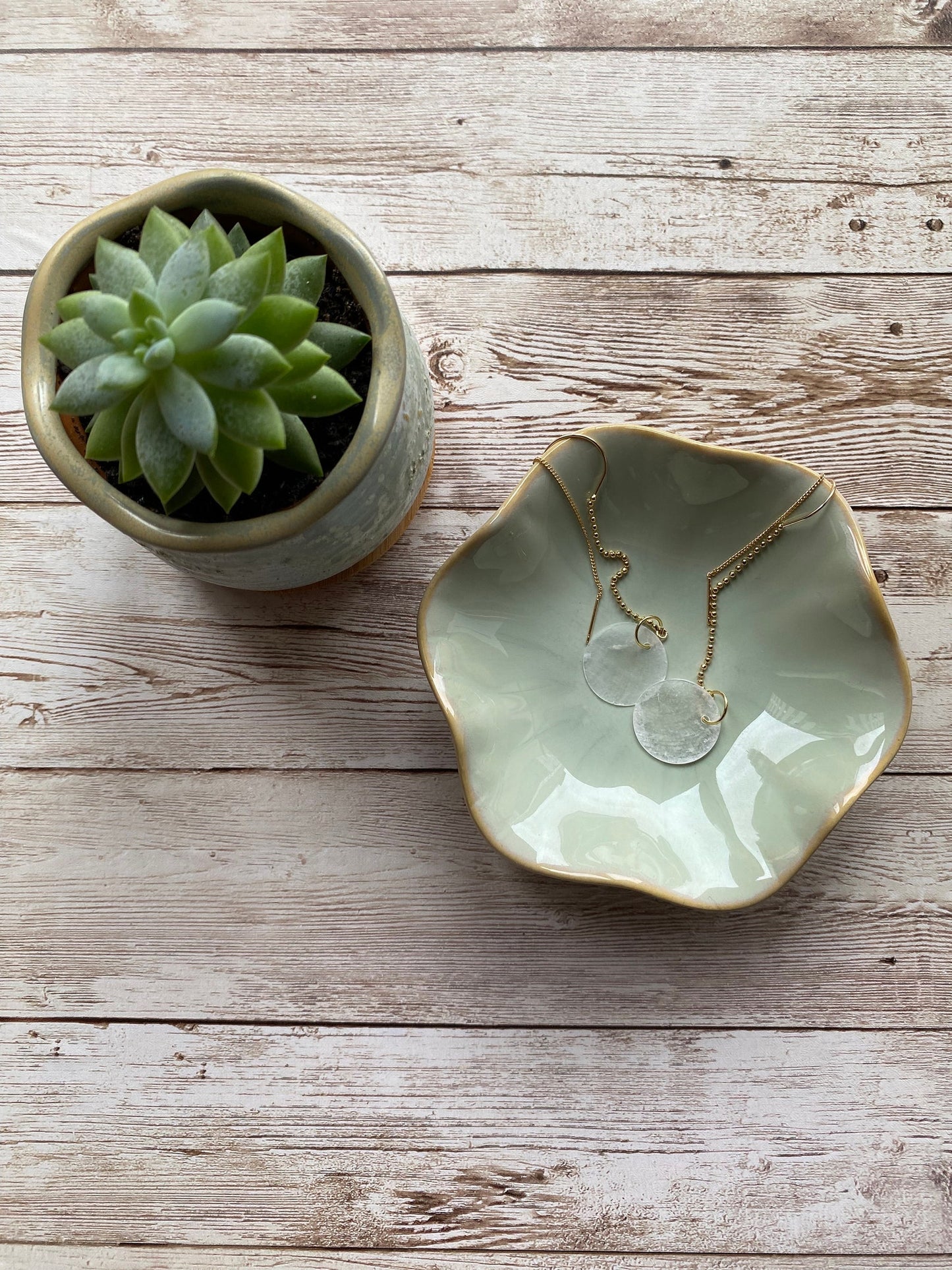 A pair of Aking Sinta gold ball chain threader earrings with round white Capiz shell charms, draped inside a matte white ceramic bowl sitting on a rustic wooden table; the long, delicate chains hang gracefully over the bowl's edge.