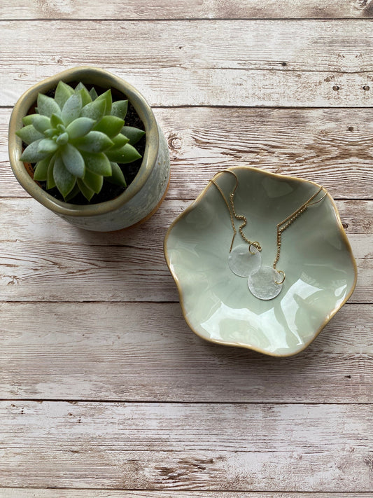 A pair of Aking Sinta gold ball chain threader earrings with round white Capiz shell charms, draped inside a matte white ceramic bowl sitting on a rustic wooden table; the long, delicate chains hang gracefully over the bowl's edge.