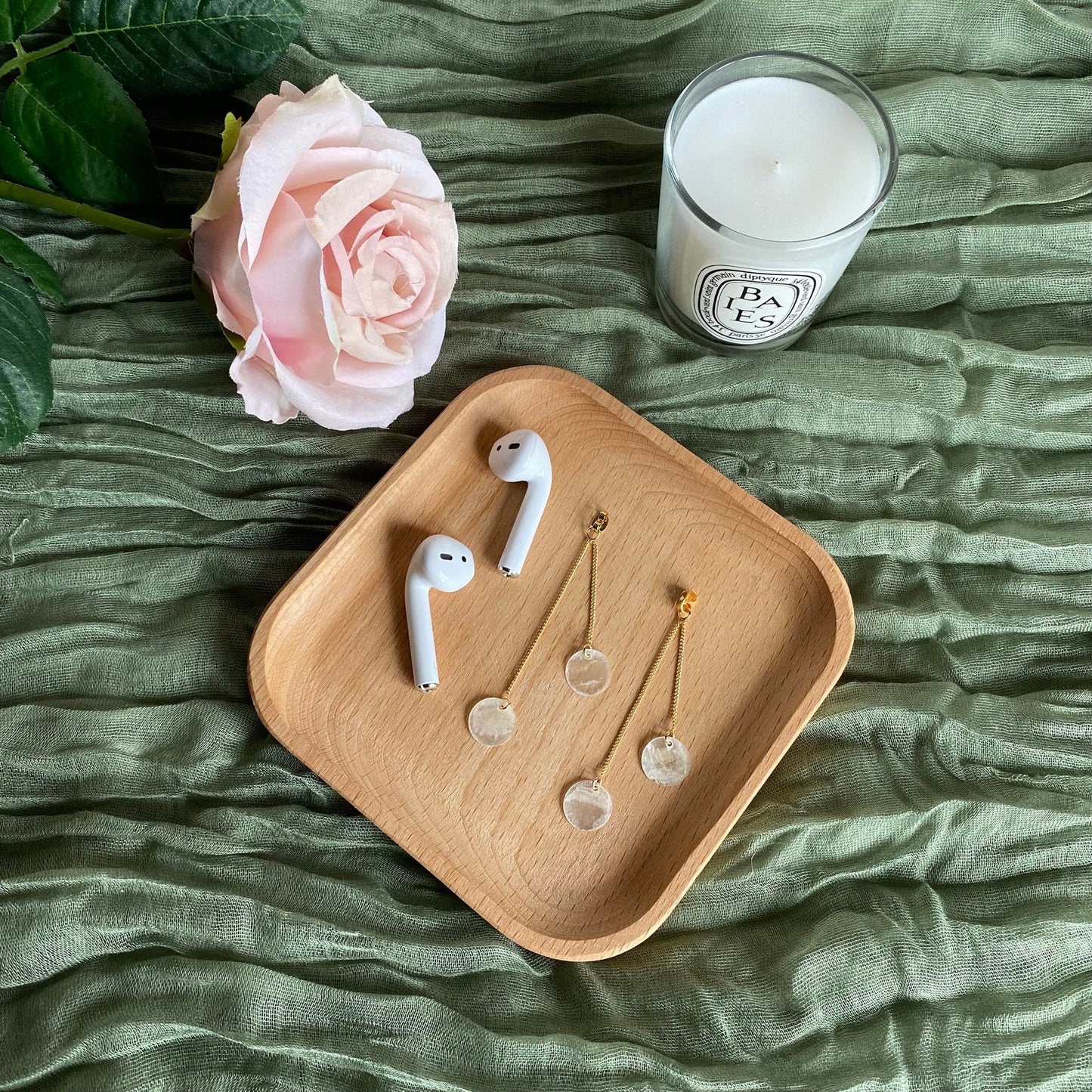 A top-down lifestyle shot of Aking Sinta 2-in-1 Capiz Shell earrings placed inside a textured wooden bowl; a pair of white earphones rests at the top of the frame, and a soft white rose is visible in the top left corner.