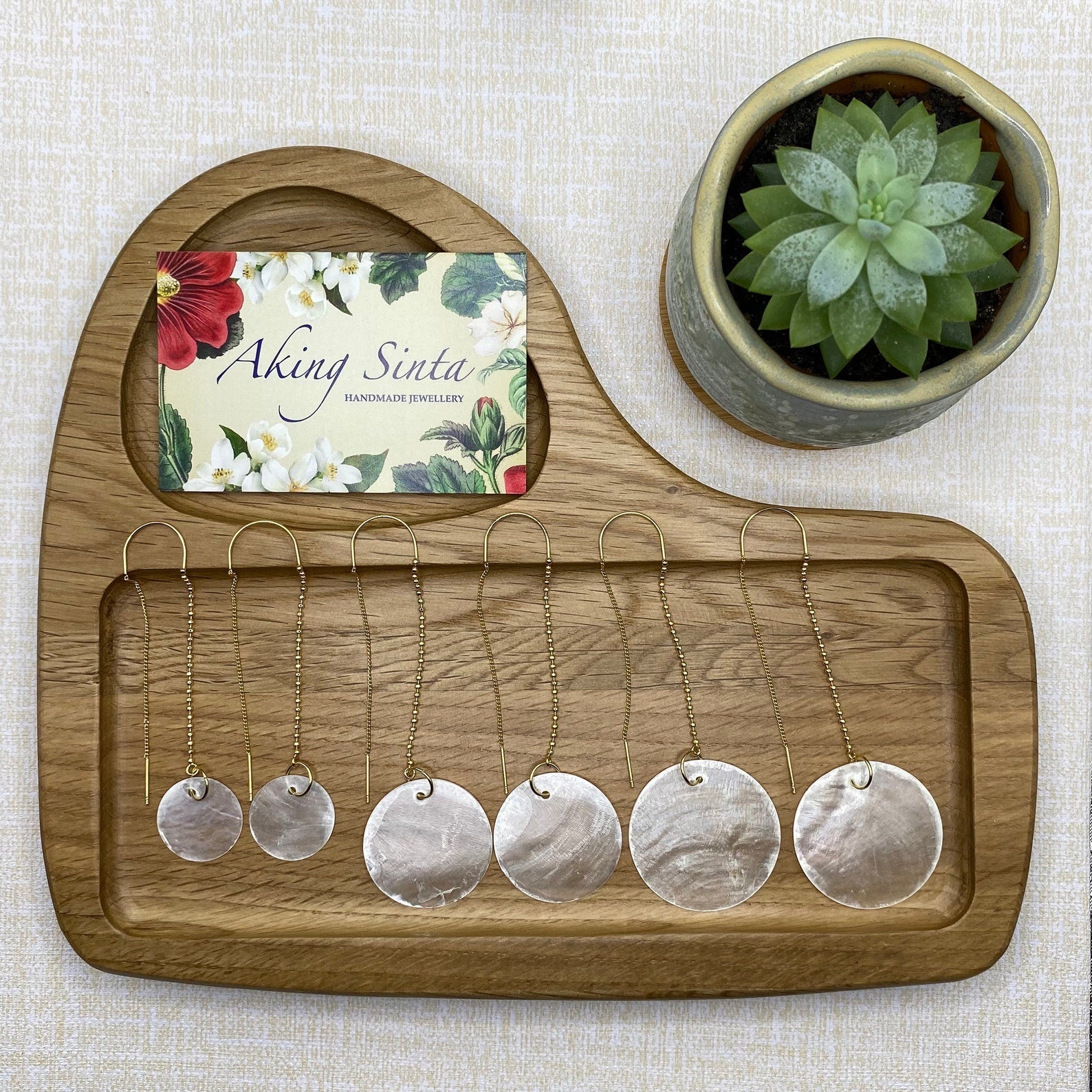 A collection of Aking Sinta round Capiz shell earrings arranged vertically on a wooden jewelry display; the sizes range from a small dainty disk at the top to a medium-sized hoop, ending with a large statement wedding-sized disk at the bottom.