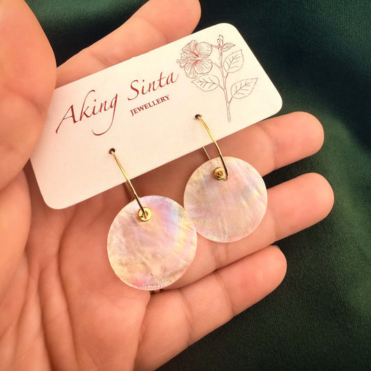 A hand holding a white display card featuring Capiz shell hoop earrings with metal beads against a dark green background.