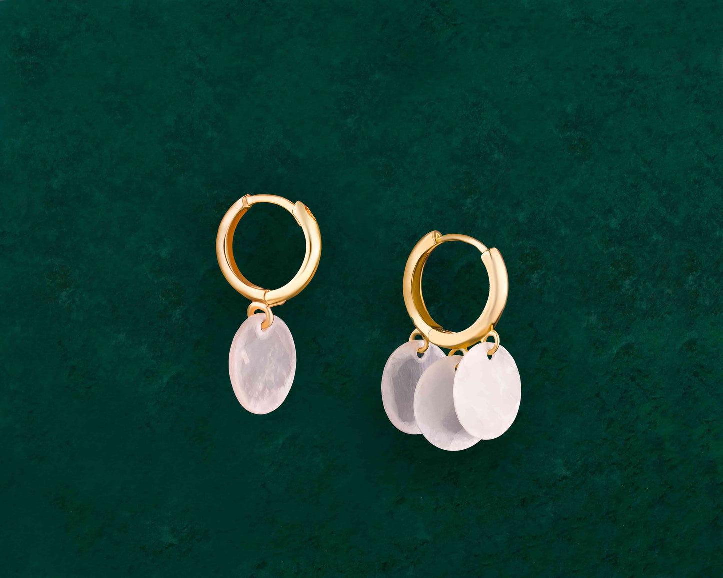 A pair of Aking Sinta gold huggie earrings displayed on a dark green background; the left earring features a single round iridescent Capiz shell charm, while the right earring features three round Capiz shell charms stacked vertically; both hang from polished gold hoops