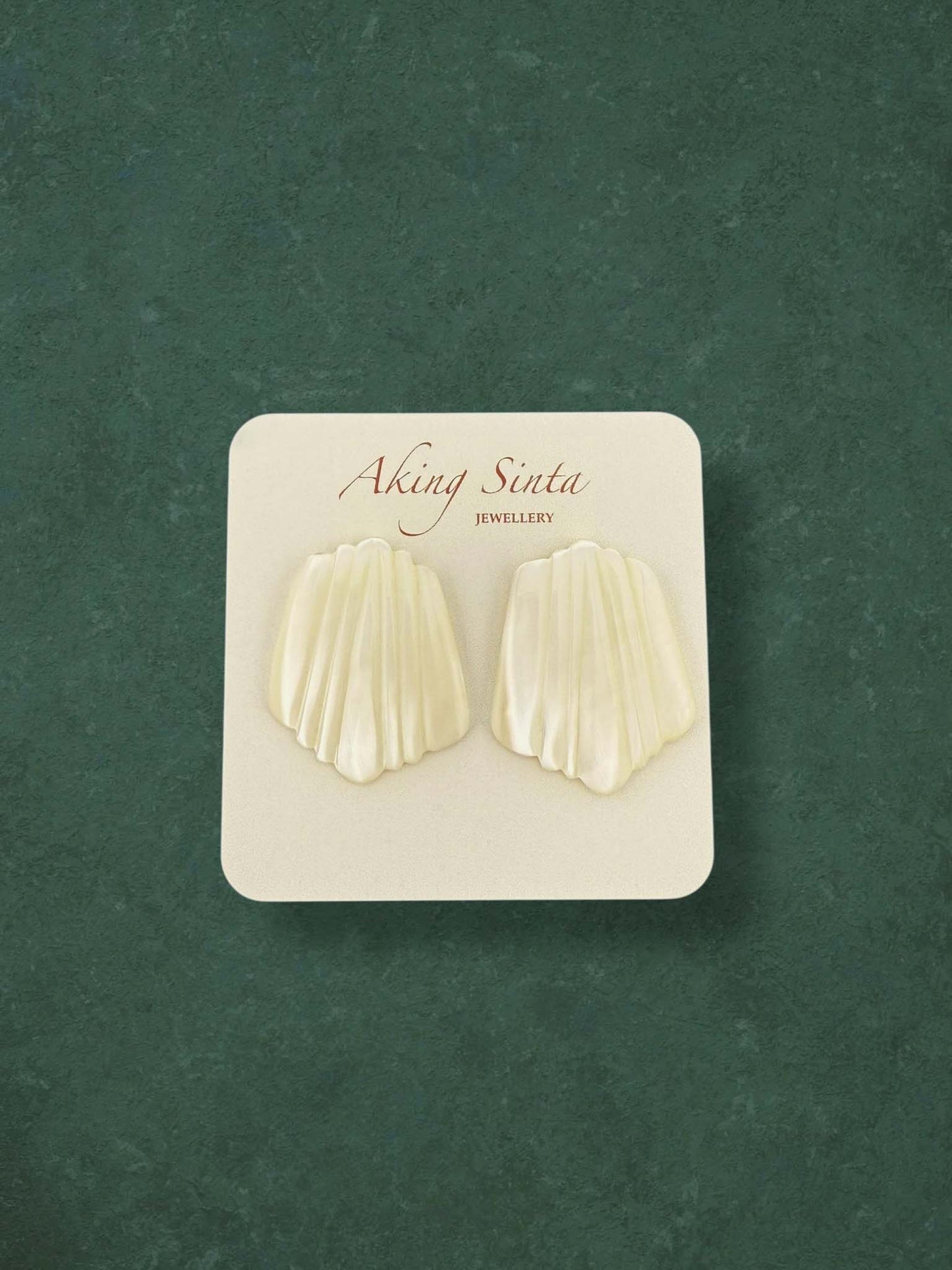 A pair of Aking Sinta Mother of Pearl stud earrings carved into a ribbed scallop shell shape. These elegant cream-colored iridescent earrings are presented on a branded white card against a green background.