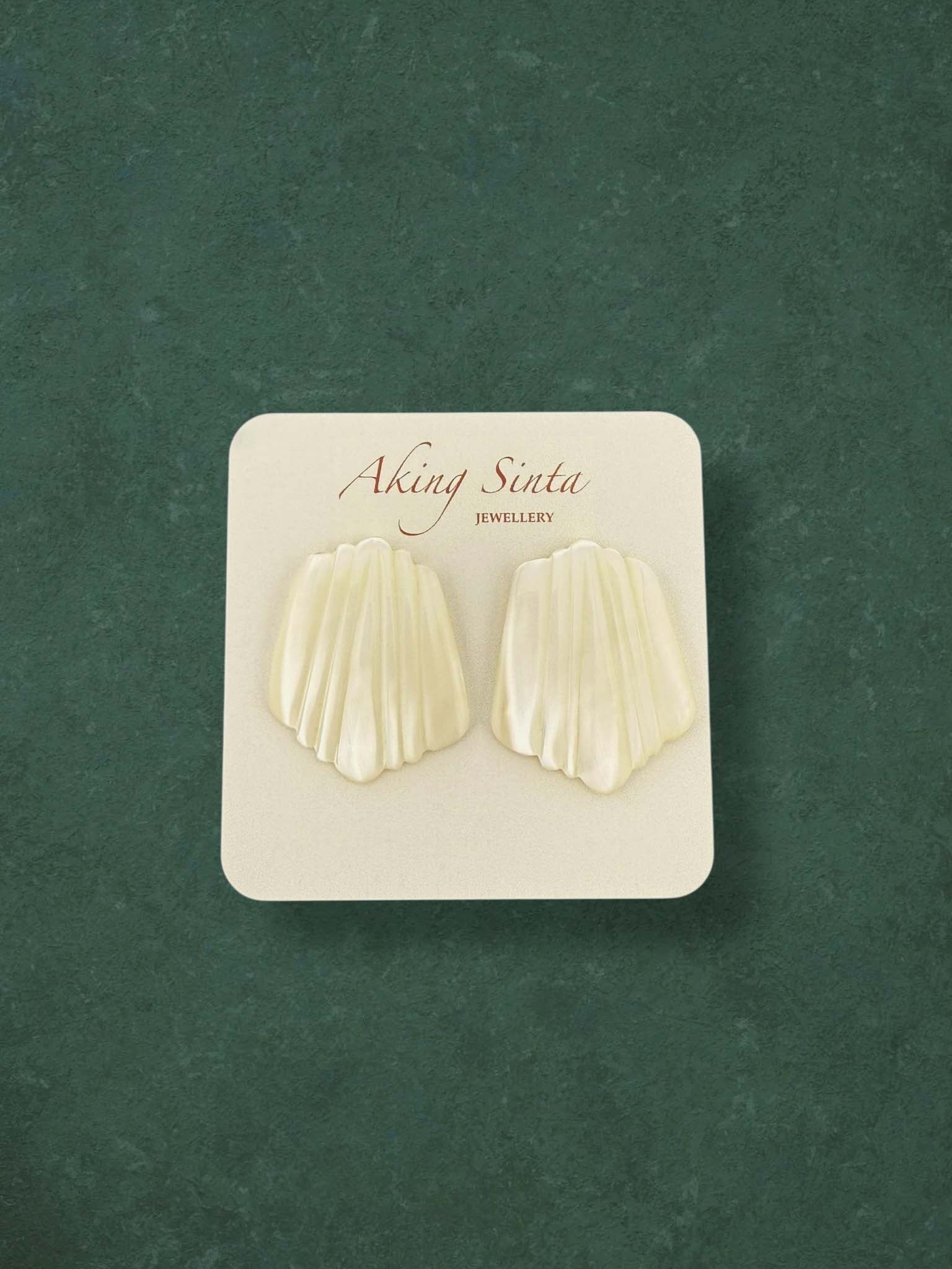 A pair of Aking Sinta Mother of Pearl stud earrings carved into a ribbed scallop shell shape. These elegant cream-colored iridescent earrings are presented on a branded white card against a green background.