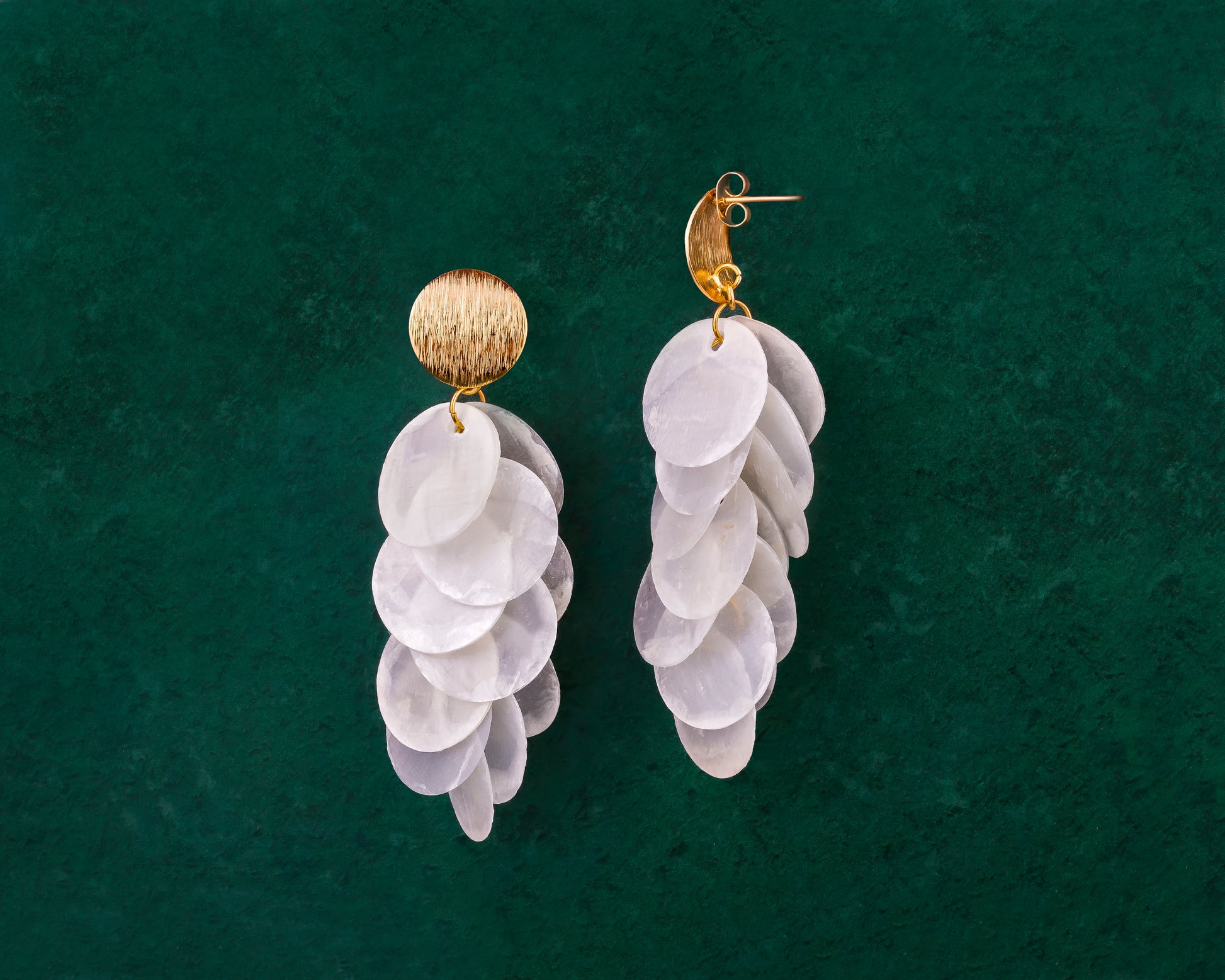 A pair of Aking Sinta gold cascading earrings on a dark green background; featuring a modern brushed-finish gold stud from which multiple iridescent white Capiz shell disks hang in a vertical, tiered sequence