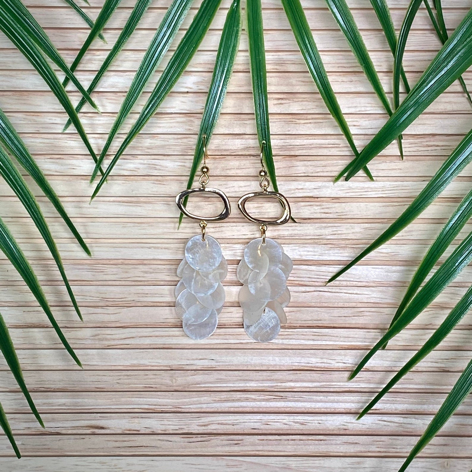 A pair of Aking Sinta Cascading Capiz Shell Earrings displayed on a warm-toned wooden surface; the iridescent white shell tiers are surrounded by vibrant green palm leaves that curve around the jewelry in a lush, tropical composition.