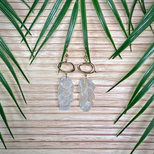 A pair of Aking Sinta Cascading Capiz Shell Earrings displayed on a warm-toned wooden surface; the iridescent white shell tiers are surrounded by vibrant green palm leaves that curve around the jewelry in a lush, tropical composition.