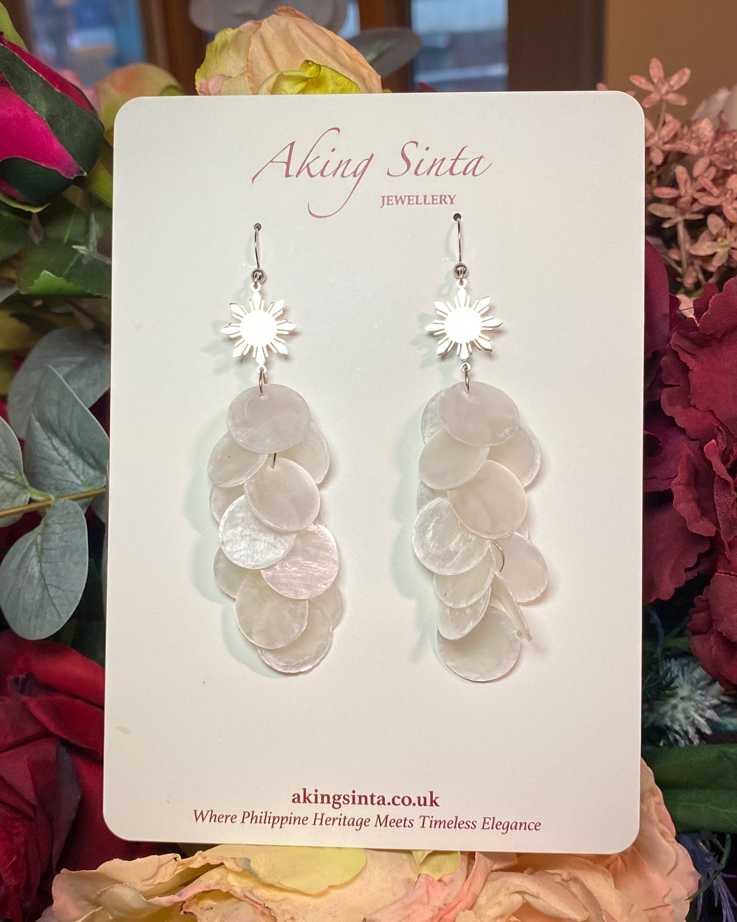 Aking Sinta Cascading Capiz Shell Hook Earrings with Araw sun charms displayed on cardboard holder with floral flowers in the background, highlighting lightweight design and Philippine-inspired craftsmanship