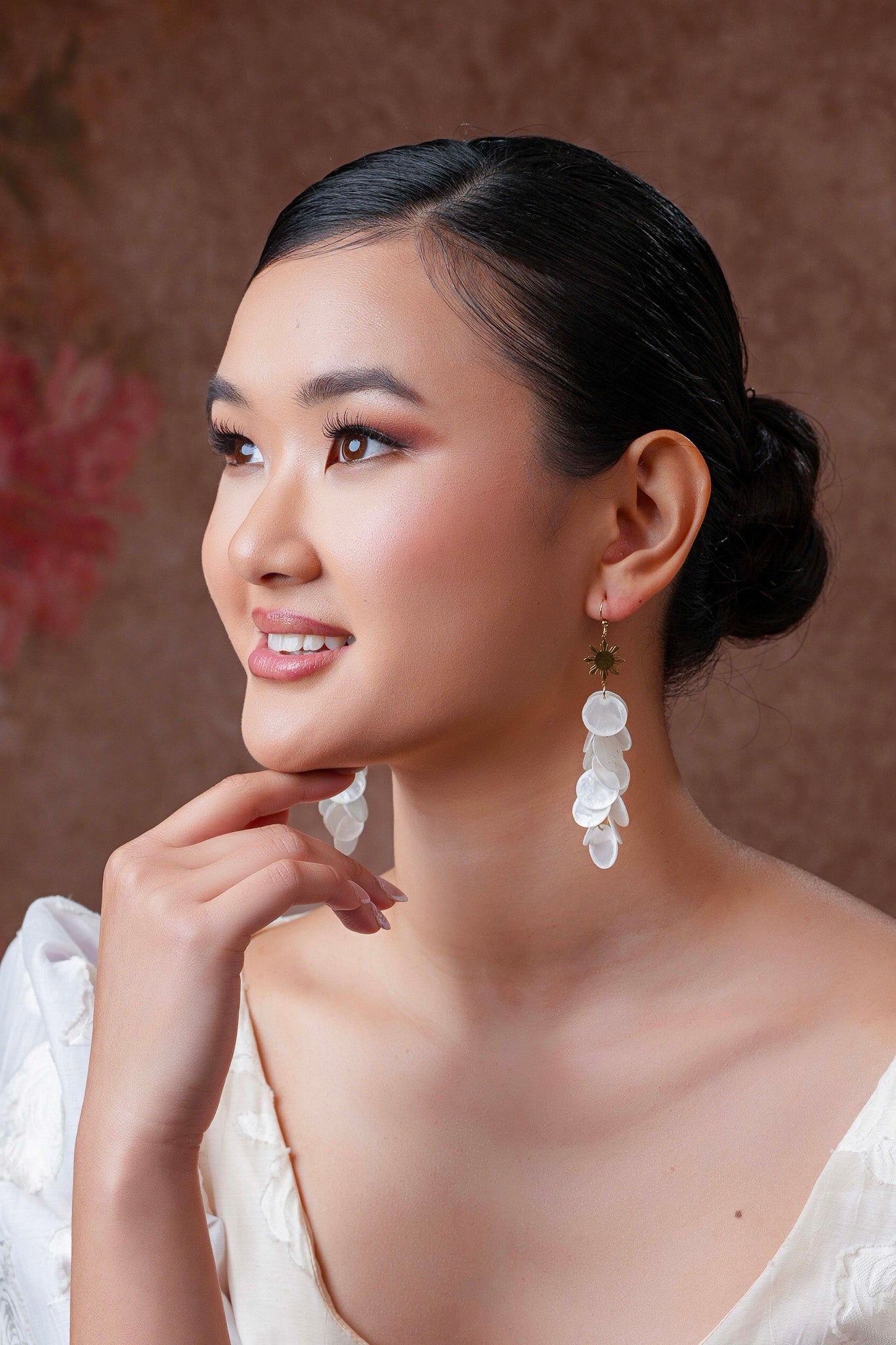 Aking Sinta Cascading Capiz Shell Hook Earrings with Araw sun charms worn by model in white, left hand on chin, looking slightly left, highlighting lightweight design and Philippine-inspired craftsmanship