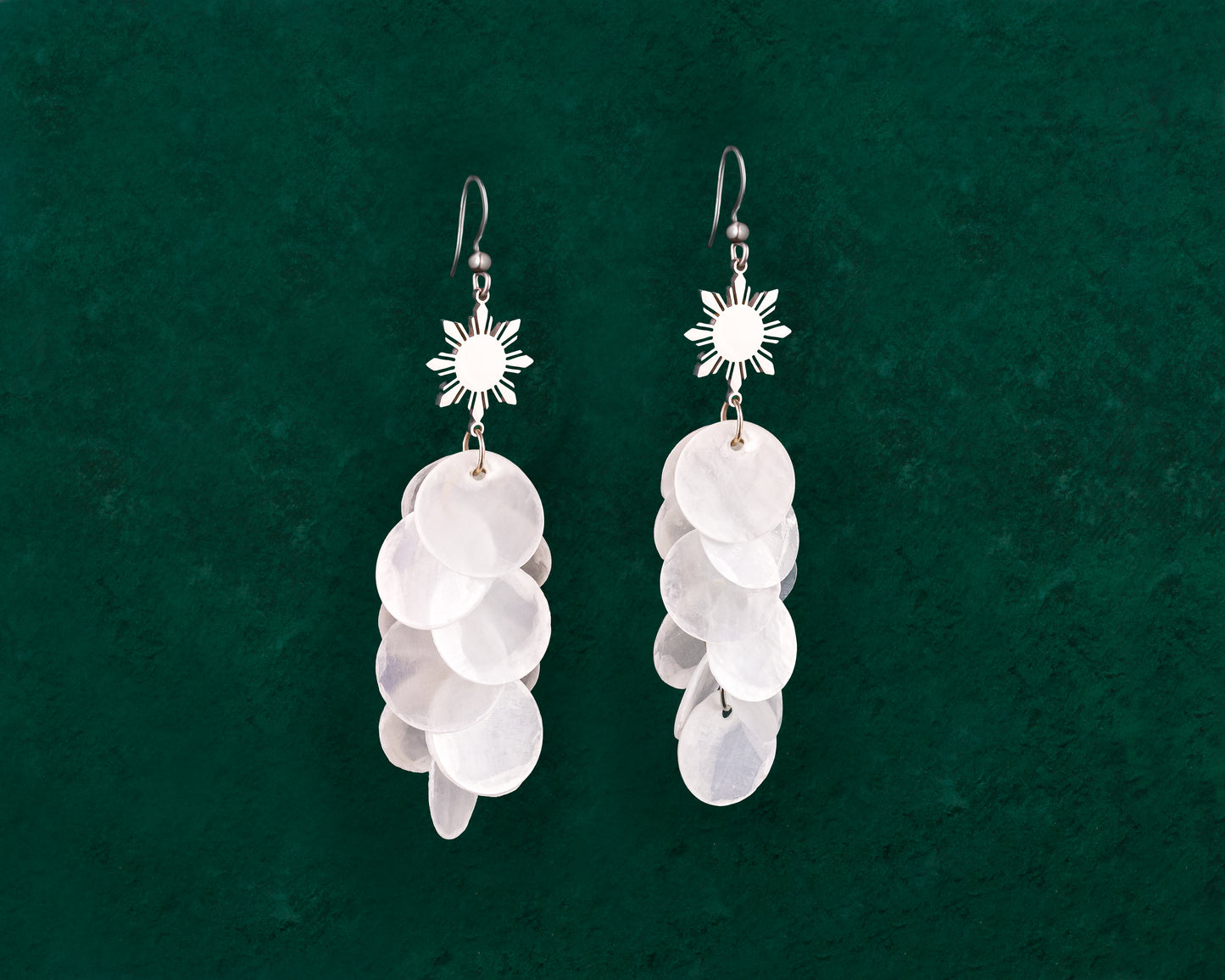 Aking Sinta Cascading Capiz Shell Hook Earrings silver variant with Araw sun charms displayed on dark green background, highlighting lightweight design and Philippine-inspired craftsmanship