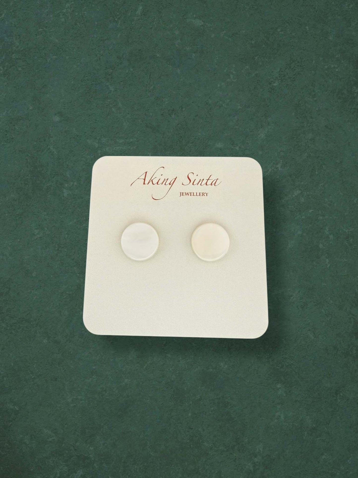A pair of Aking Sinta classic round mother of pearl stud earrings. These minimalist circular shell earrings feature a smooth, iridescent white finish and are displayed on a branded white card