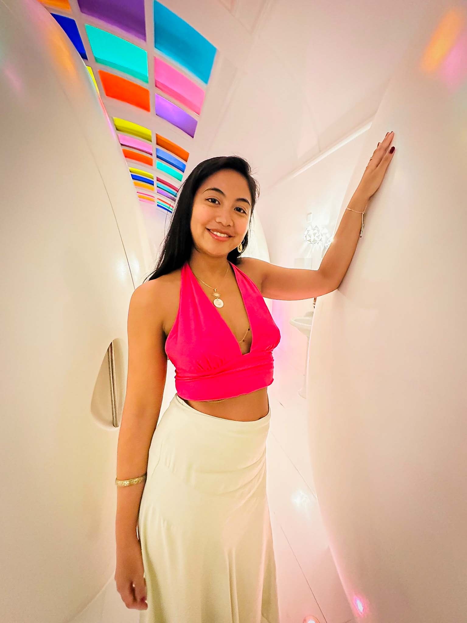 A full-body shot of a smiling woman posing in a modern, brightly lit corridor with colorful ceiling accents. She is wearing a vibrant pink halter top and a long skirt, accessorized with an Aking Sinta gold sun-motif and Capiz shell pendant necklace. The image showcases how the jewelry complements a chic, contemporary outfit.