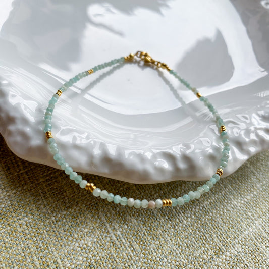 A dainty Aking Sinta aquamarine anklet with small gold bead accents, resting inside an iridescent mother-of-pearl shell container; the light blue stones contrast beautifully with the shimmering, pearlescent background.