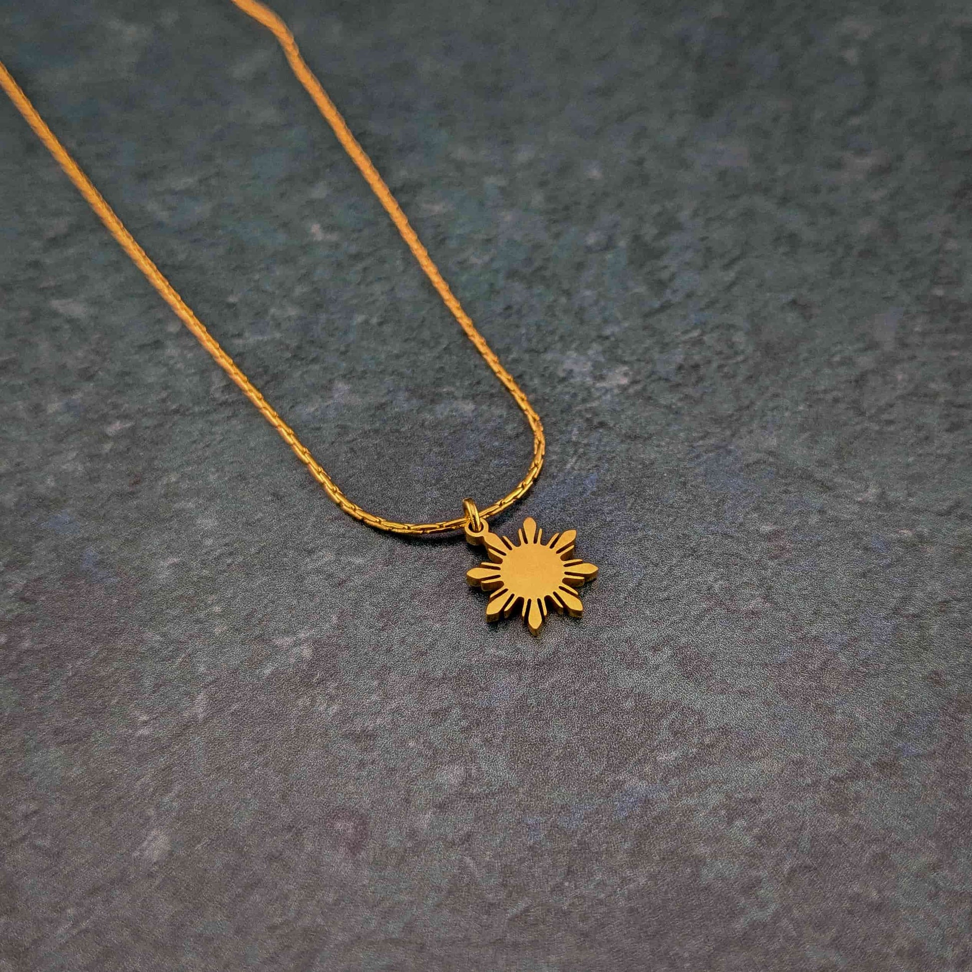 Aking Sinta Dainty Araw Necklace in gold displayed on rock-like textured background, showcasing 8-ray sun design, Philippine-inspired jewellery with subtle heritage