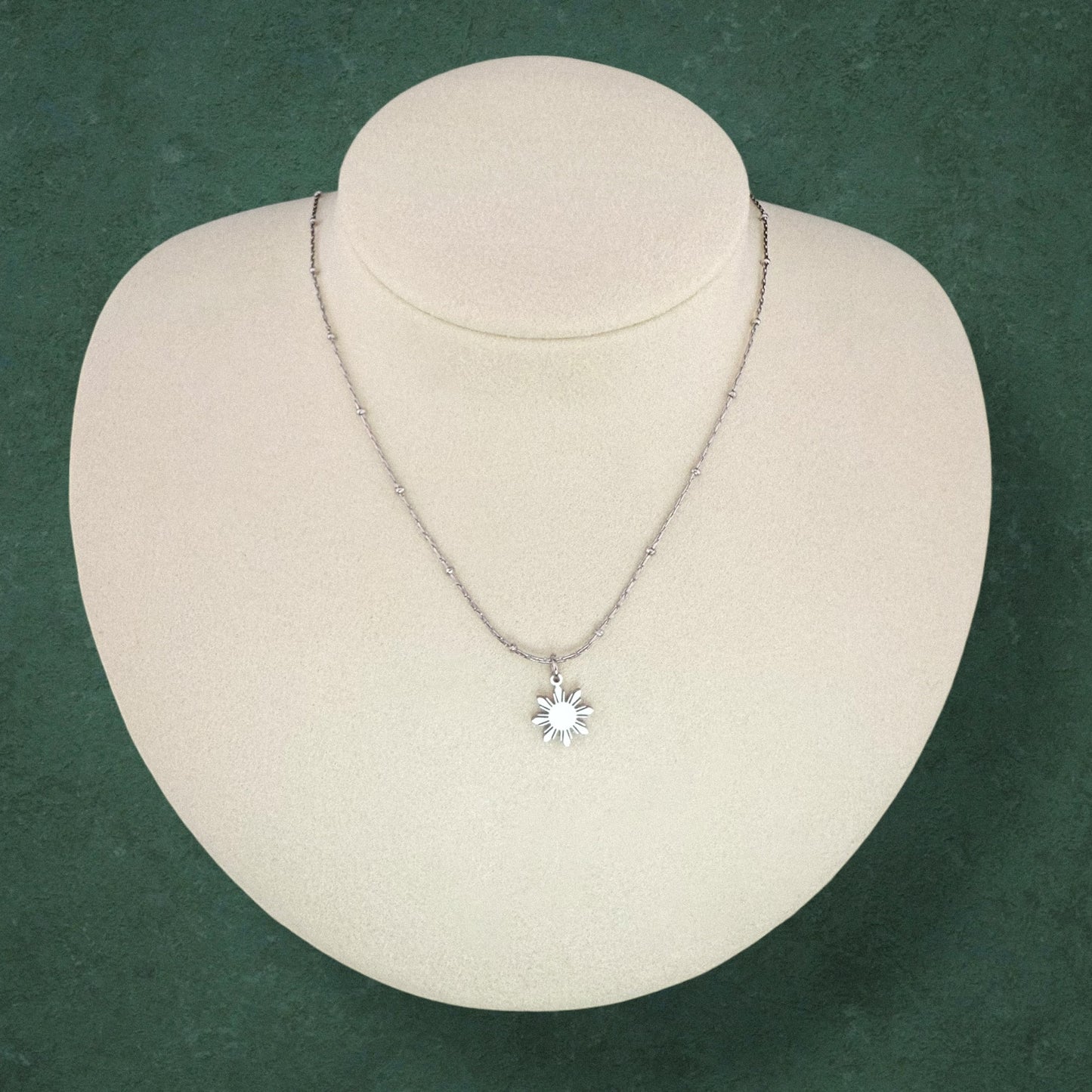Aking Sinta Dainty Araw Necklace in silver displayed on necklace holder on dark green background, showcasing 8-ray sun design, Philippine-inspired jewellery with subtle heritage