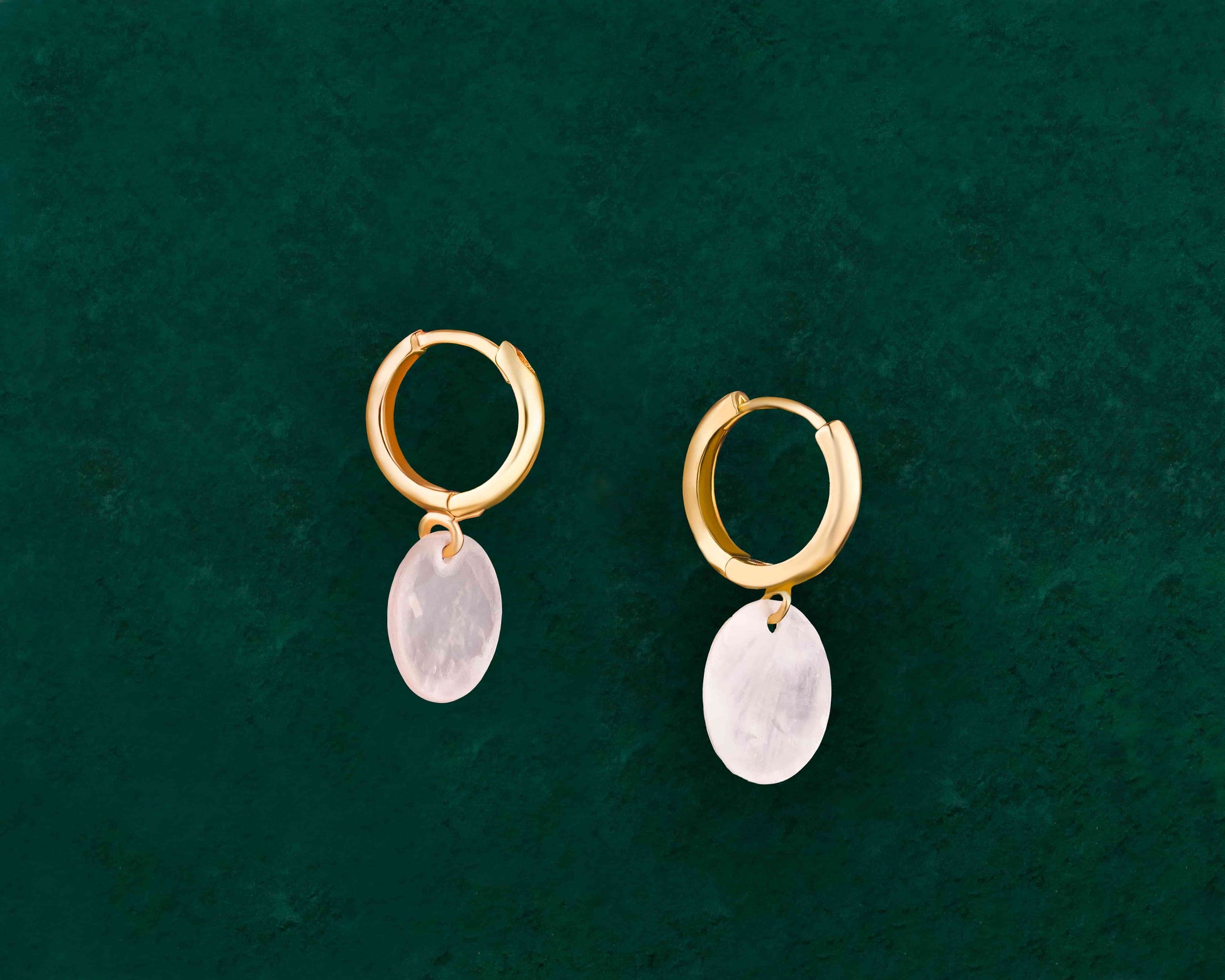 A pair of Aking Sinta Dainty Capiz Shell Huggie Earrings in gold, displayed on a dark green background; featuring small, round iridescent white Capiz shell charms hanging from polished gold huggie hoops
