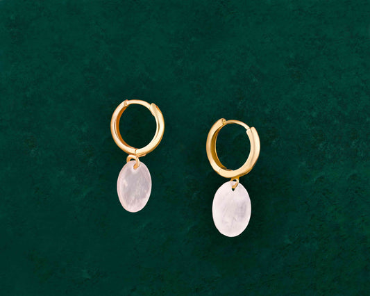 A pair of Aking Sinta Dainty Capiz Shell Huggie Earrings in gold, displayed on a dark green background; featuring small, round iridescent white Capiz shell charms hanging from polished gold huggie hoops