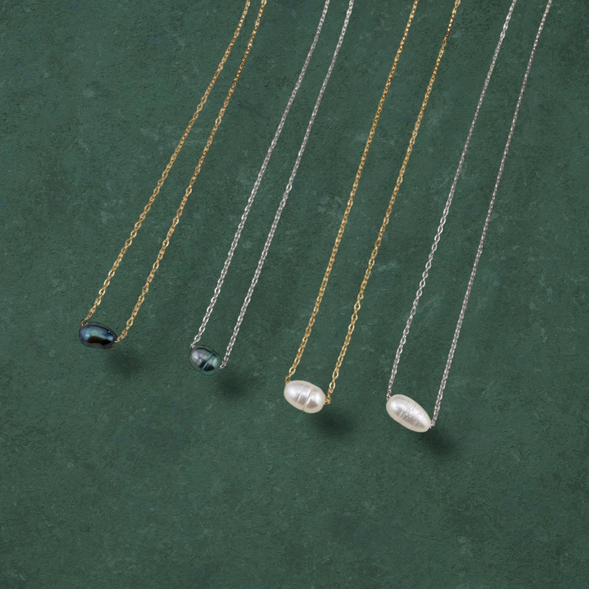 A curated flat-lay of Aking Sinta floating pearl necklaces, showcasing four variations featuring lustrous white and iridescent dark pearls strung on delicate gold and silver chains. The necklaces are elegantly arranged in parallel on a rich, deep green textured background, highlighting the minimalist and versatile design of the collection