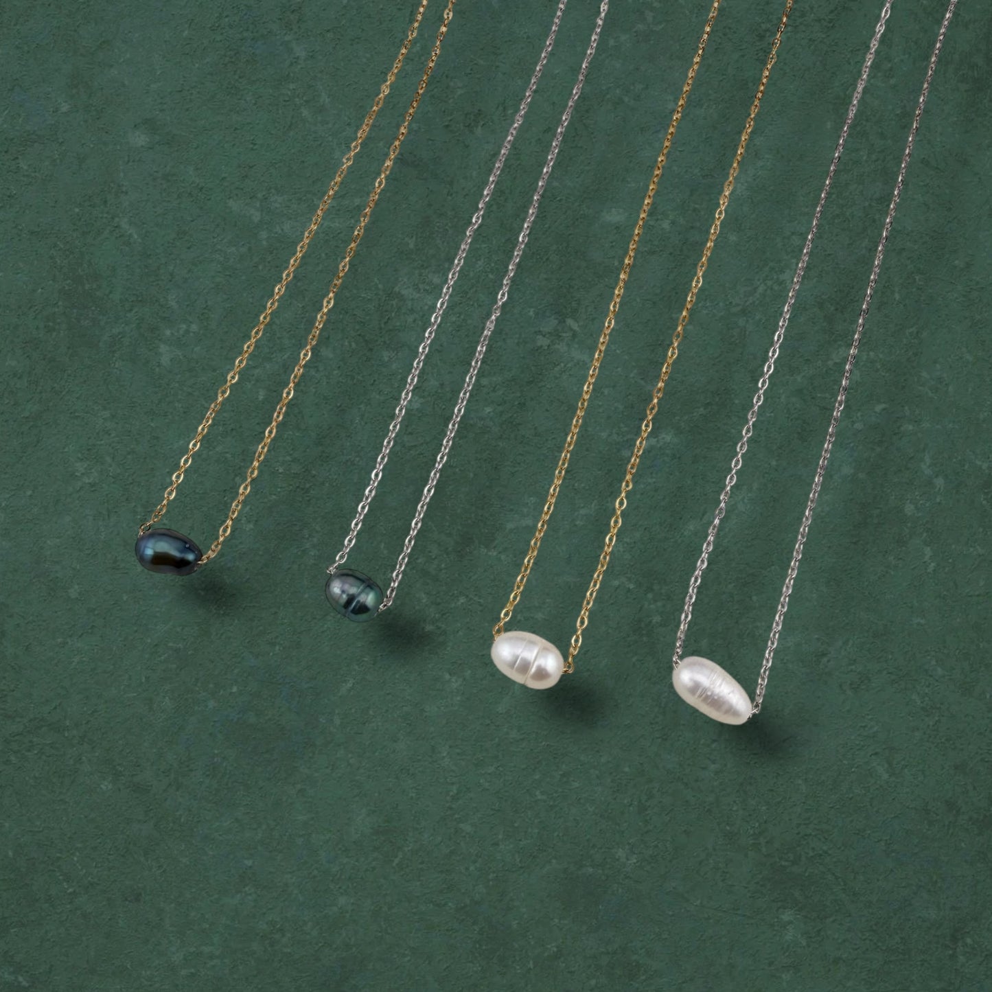 A curated flat-lay of Aking Sinta floating pearl necklaces, featuring four variations with lustrous white and iridescent dark pearls suspended on delicate gold and silver chains. The necklaces are elegantly arranged in parallel on a rich, deep green textured background, highlighting the minimalist and versatile design of the collection