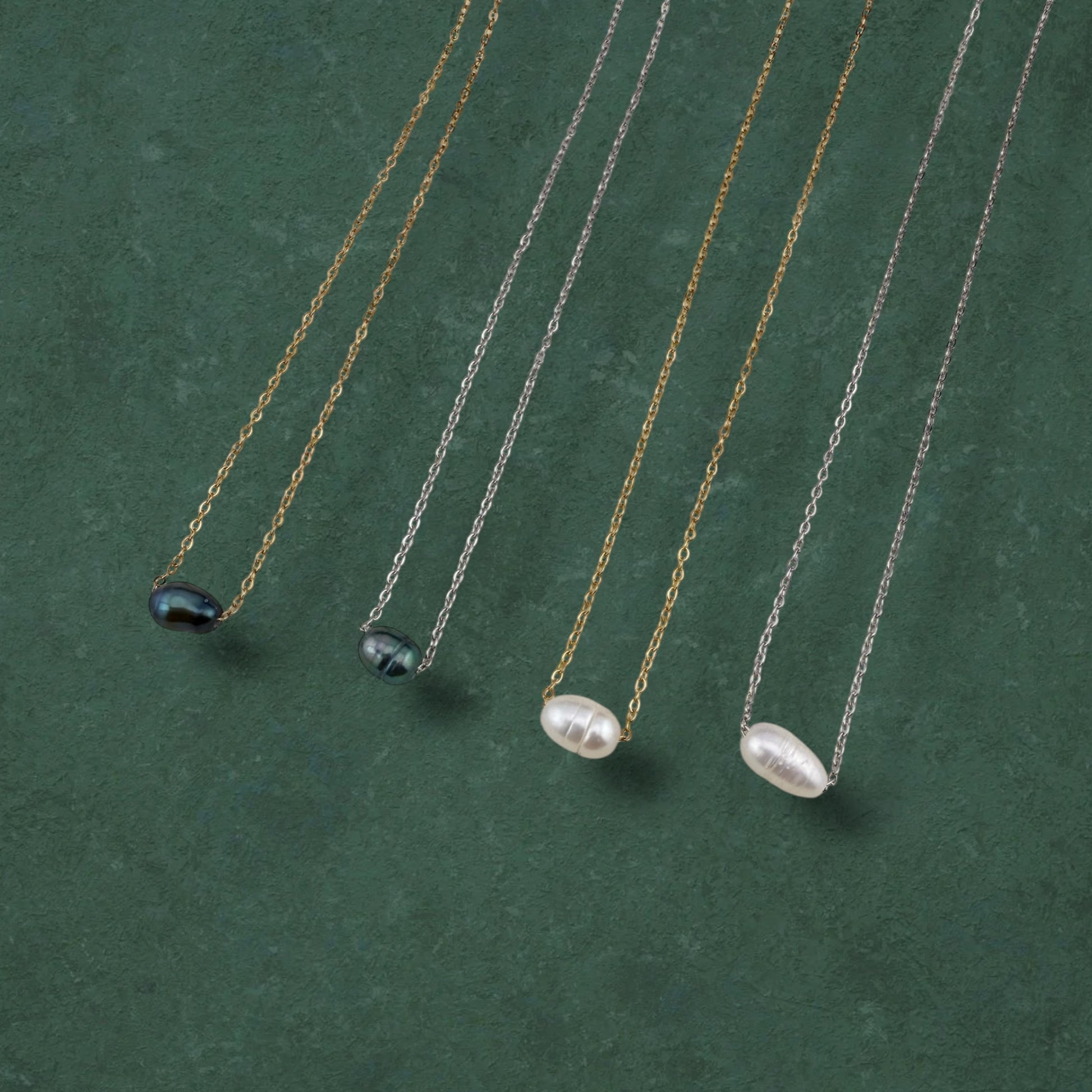 A curated flat-lay of Aking Sinta floating pearl necklaces, featuring four variations with lustrous white and iridescent dark pearls suspended on delicate gold and silver chains. The necklaces are elegantly arranged in parallel on a rich, deep green textured background, highlighting the minimalist and versatile design of the collection