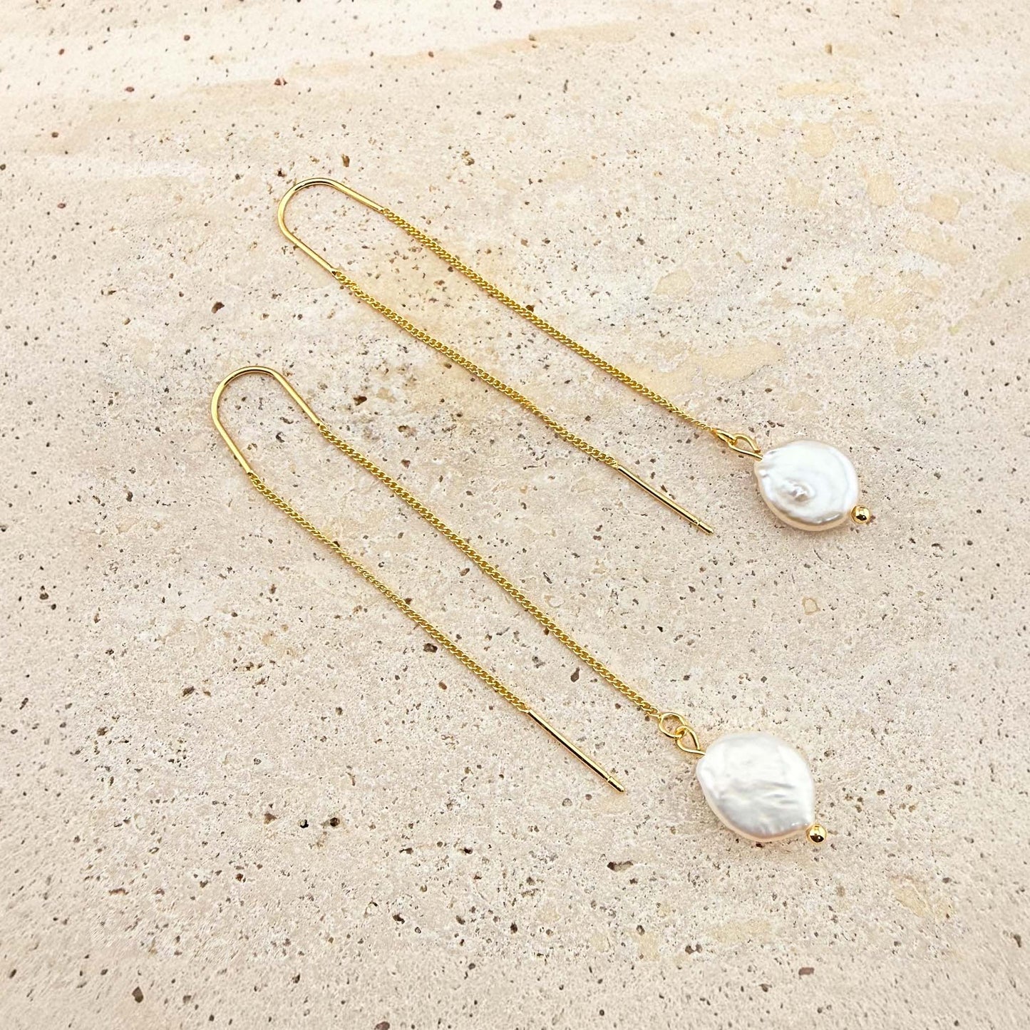 A pair of elegant Aking Sinta gold chain threader earrings, each featuring a single, lustrous white pearl drop at the end. The delicate gold chains are laid out gracefully on a light, textured natural stone background, highlighting their minimalist and sophisticated design