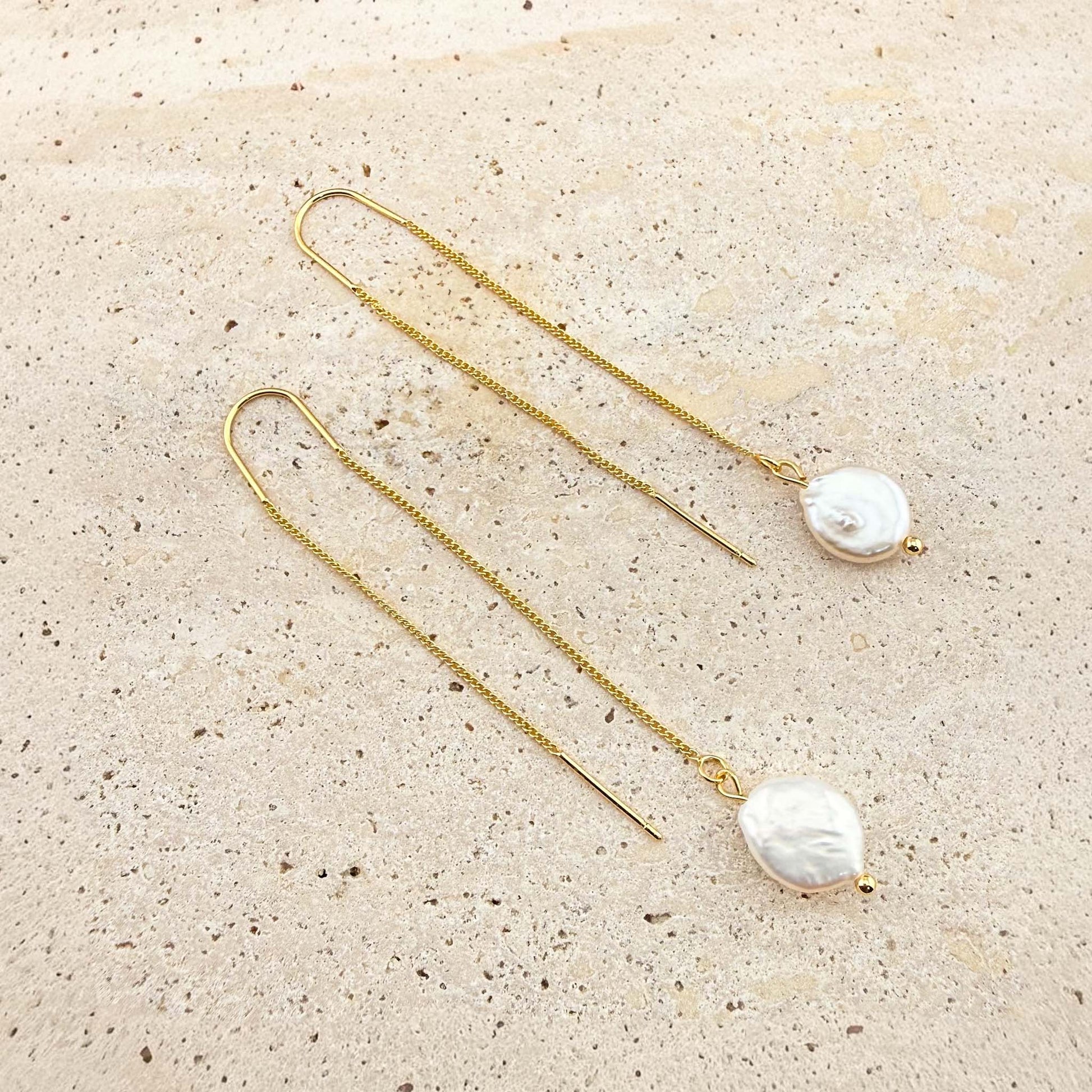 A pair of elegant Aking Sinta gold chain threader earrings, each featuring a single, lustrous white pearl drop at the end. The delicate gold chains are laid out gracefully on a light, textured natural stone background, highlighting their minimalist and sophisticated design