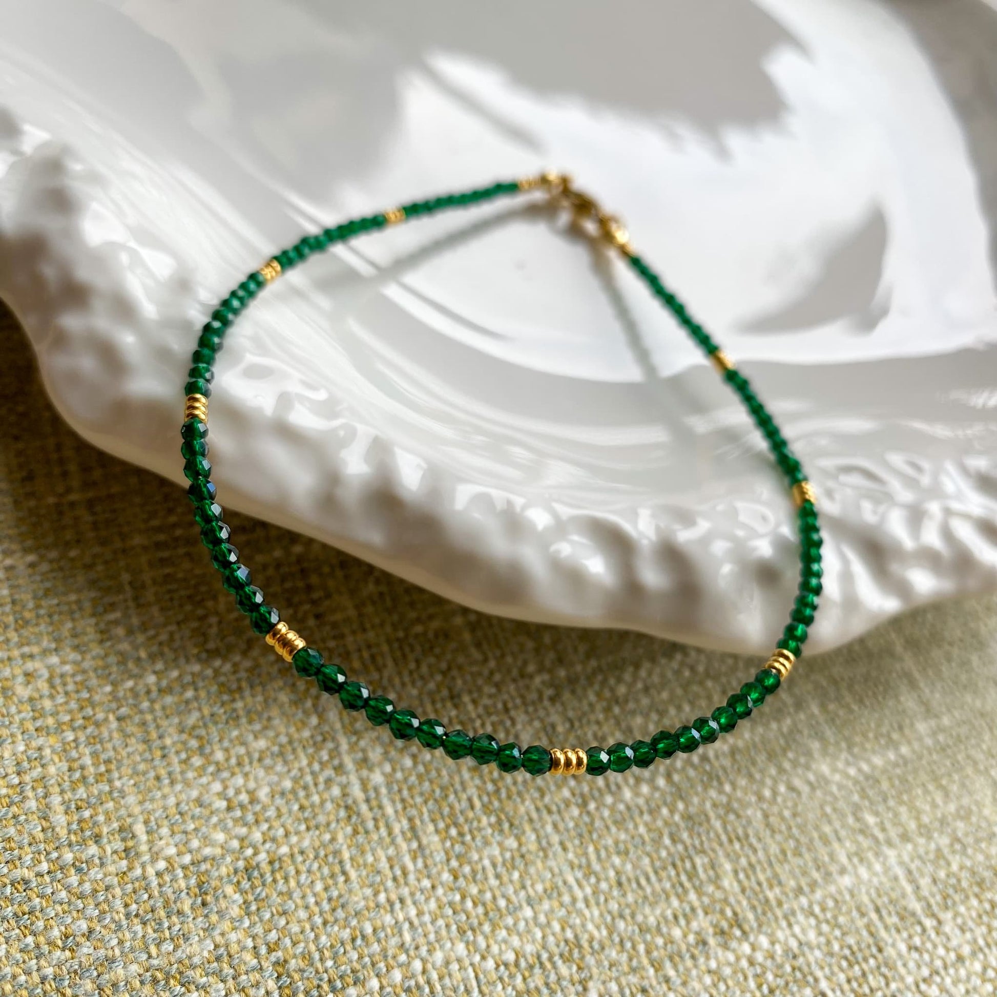 A delicate Aking Sinta gold chain anklet featuring tiny, spaced-out Green Jade beads, resting on a shimmering Mother of Pearl shell dish that reflects iridescent pink and white light.