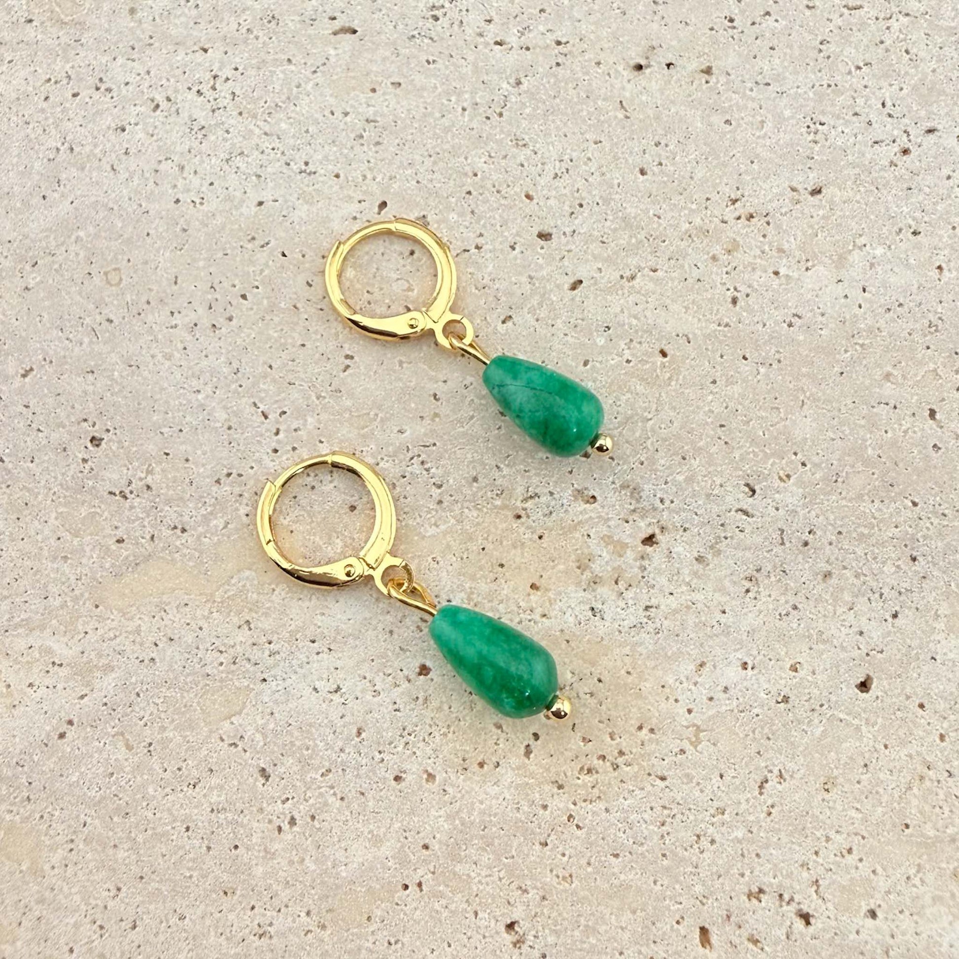 A pair of Aking Sinta gold huggie hoop earrings featuring polished green jade teardrop pendants. These dainty gemstone earrings are shown on a natural sand-colored stone background