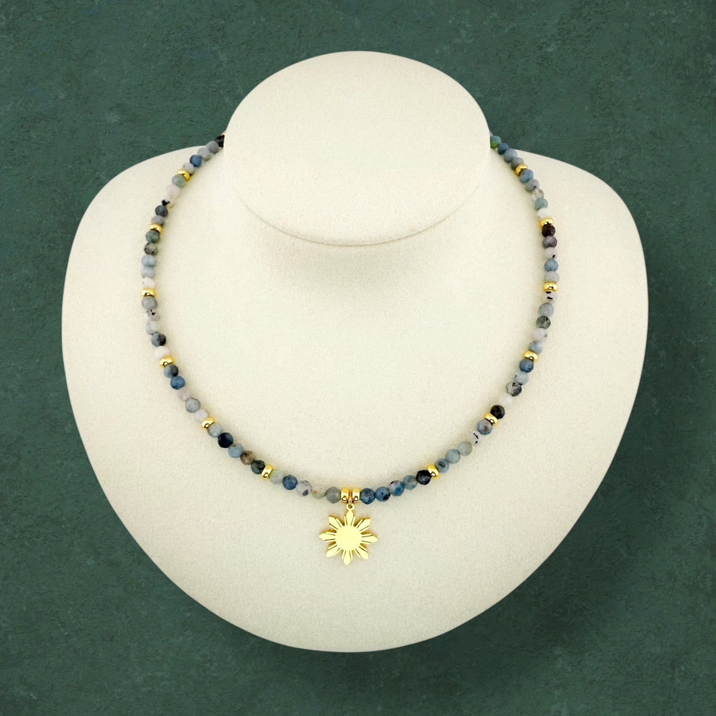 Aking Sinta Dirty Aquamarine necklace on a white display bust; featuring a mix of light and dark blue beads with small gold bead accents and a central 8-ray Araw sun charm.