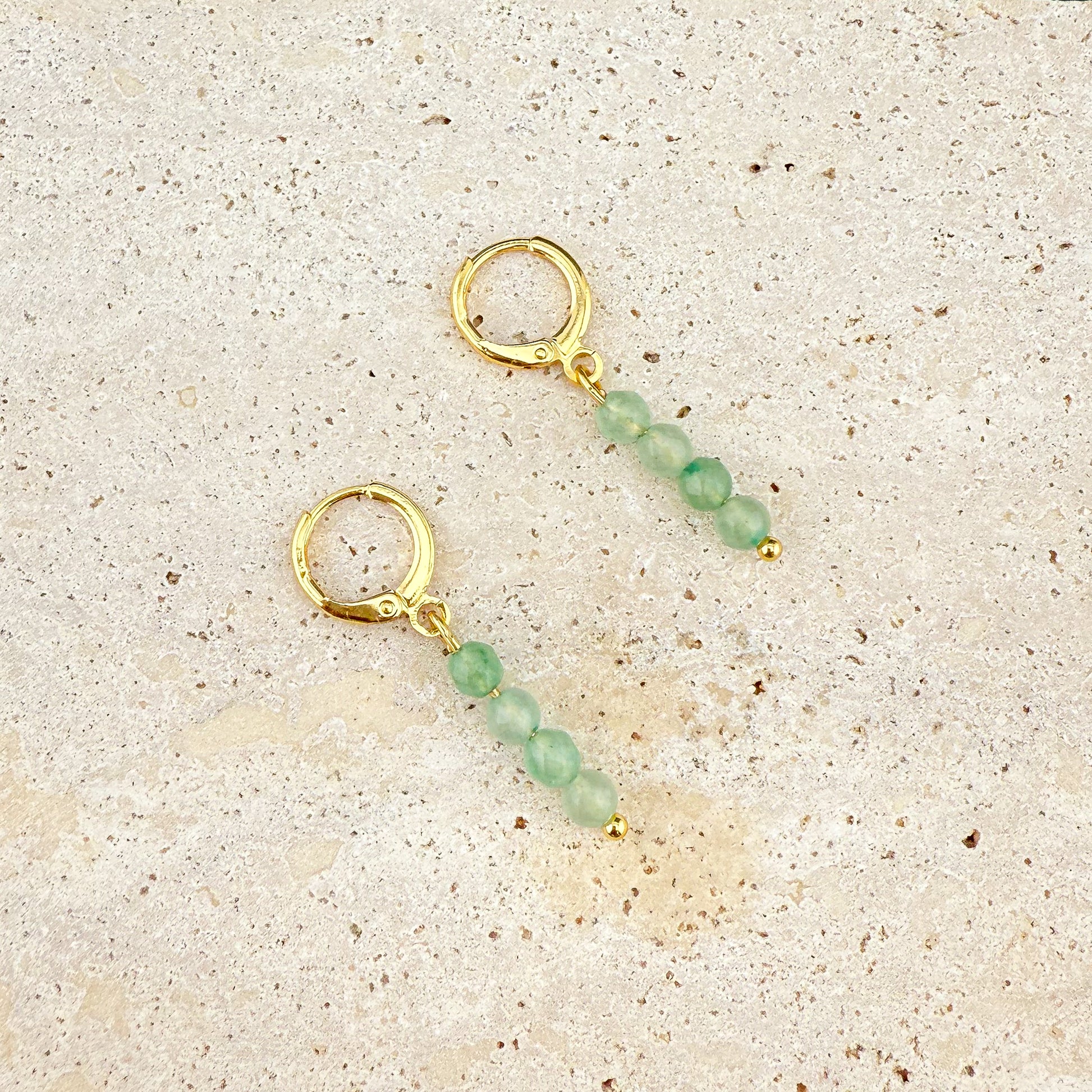 A pair of gold huggie earrings on white marble featuring faceted Green Aventurine charms; the deep green gemstones are cut with multiple light-reflecting faces, hanging from polished gold circular hoops.
