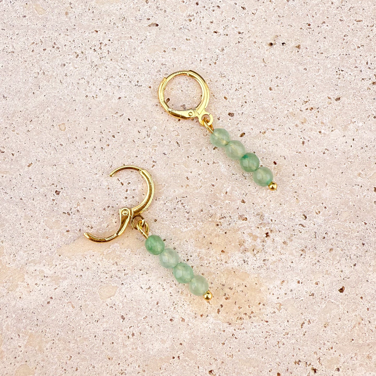 A close-up on white marble of a faceted Green Aventurine huggie earring with its gold hinge clasp open, revealing the secure snapping mechanism and the intricate light-reflecting faces of the deep green gemstone charm.