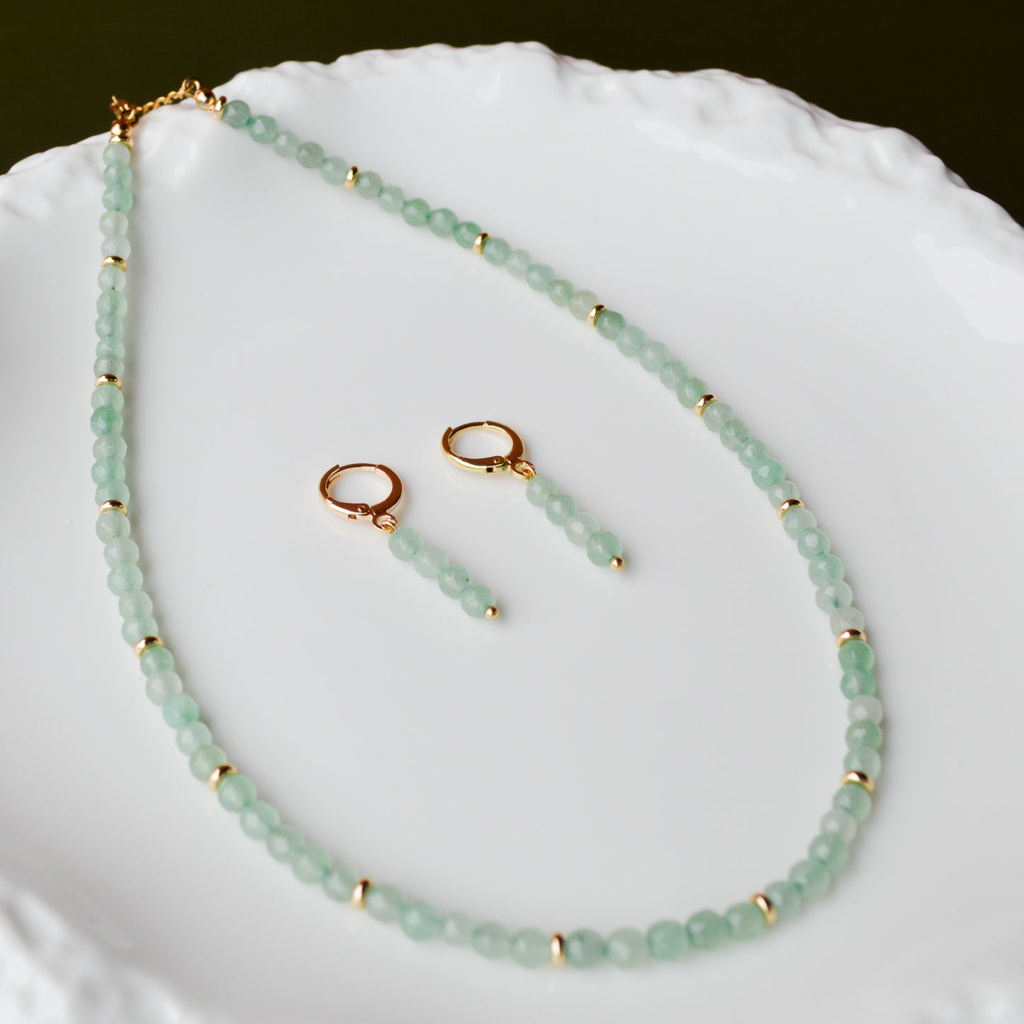 A pair of gold huggie earrings with faceted Green Aventurine charms resting on an iridescent Mother of Pearl tray; one earring is open to show the gold hinge clasp, while the iridescent shell background reflects soft pink and blue hues onto the green stones.