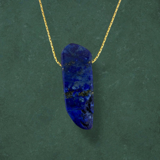 Aking Sinta floating necklace featuring a raw chunk of deep blue Lapis Lazuli with gold pyrite flecks, hanging on a delicate gold chain against a dark green background.
