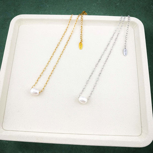 A gold and a silver Aking Sinta paperclip chain necklace, each featuring a single large white floating pearl pendant, displayed side-by-side on a white jewelry tray with a dark green background.