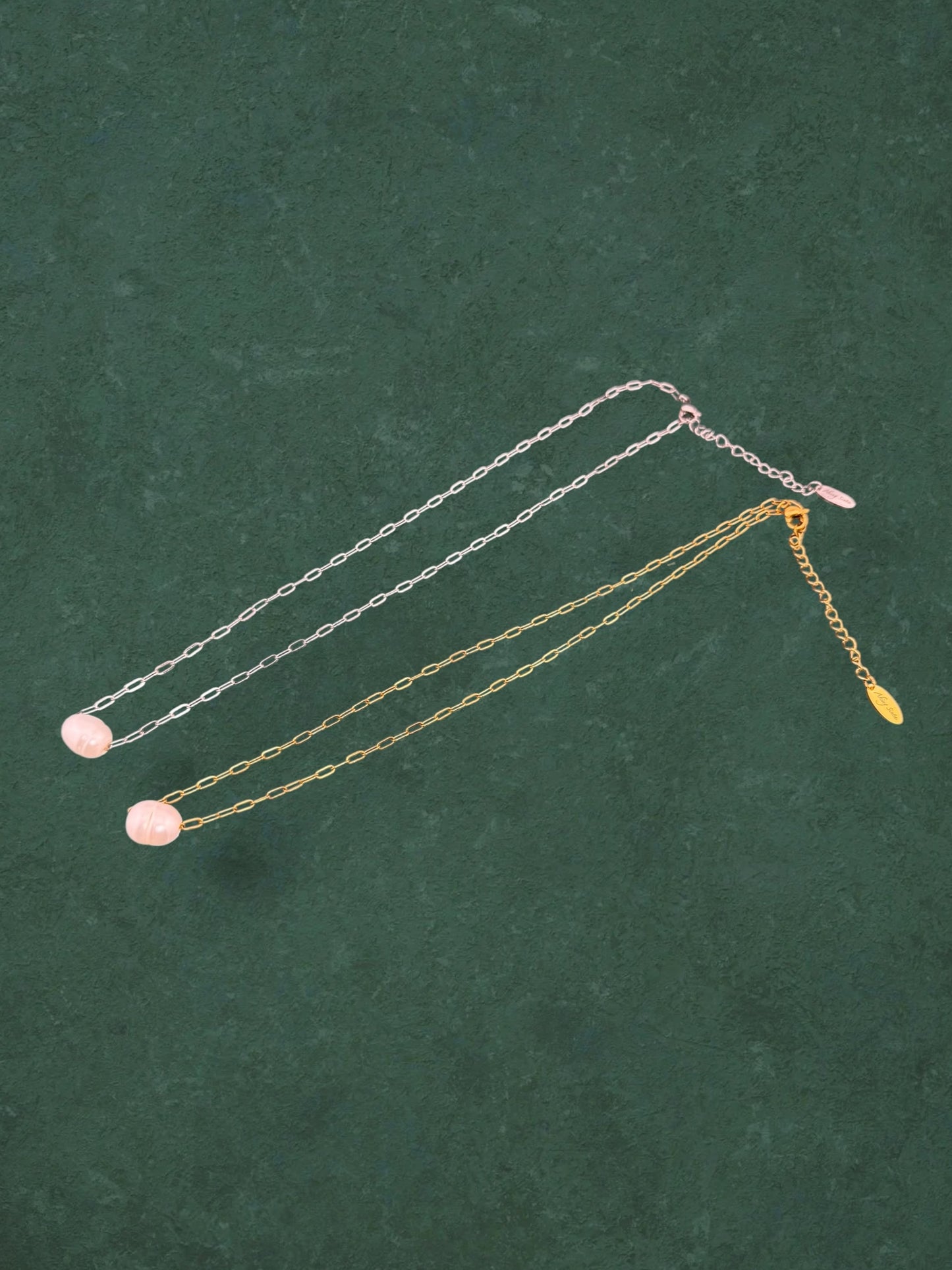 Two Aking Sinta floating pink pearl necklaces on gold and silver paperclip chains. Minimalist link jewelry with adjustable extenders and signature tags on a green background.