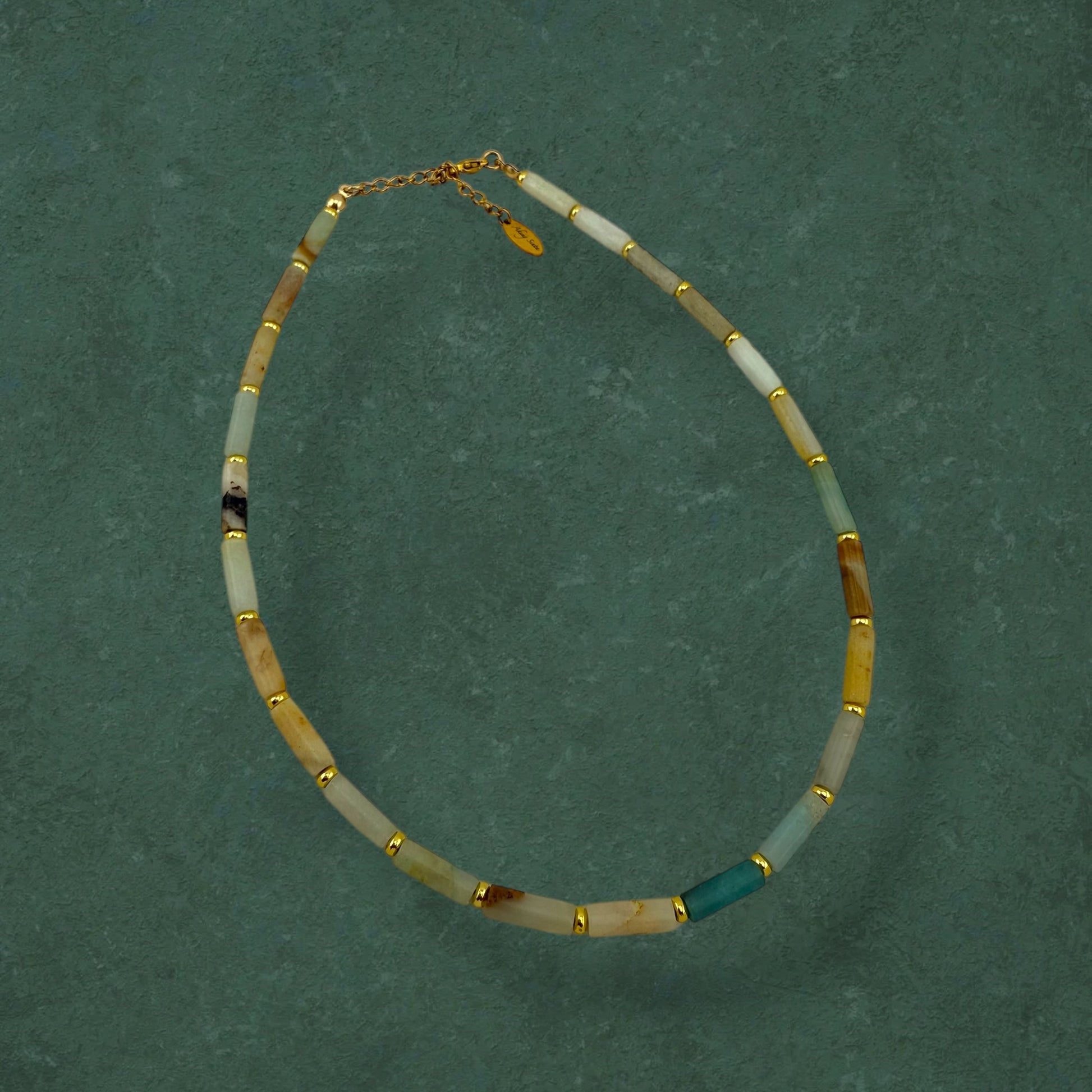 Aking Sinta Flower Amazonite necklace featuring long column-shaped beads in mint and cream, laid flat on a rich dark green background with gold bead accents.