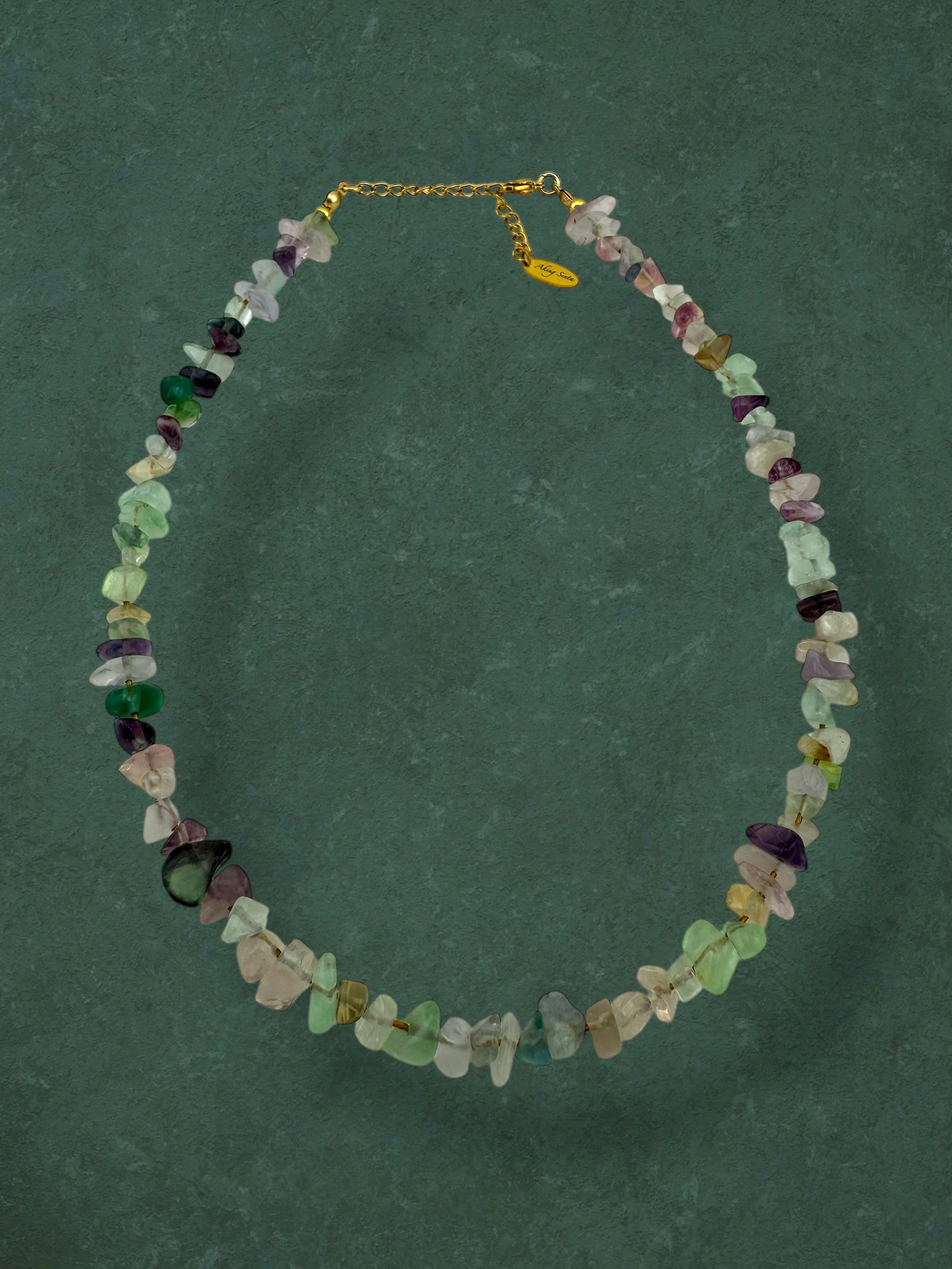 A statement Aking Sinta necklace of raw, polished Fluorite nuggets on a white neck bust; the translucent purple and green gemstones are set against a solid forest-green background.