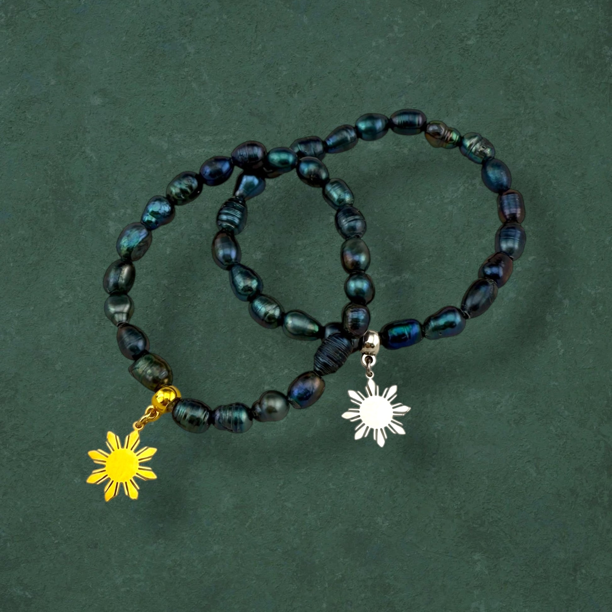 Aking Sinta Freshwater Pearl Stretch Bracelet with dark blue beads, featuring gold and silver Araw sun charms, displayed on hand against dark green background
