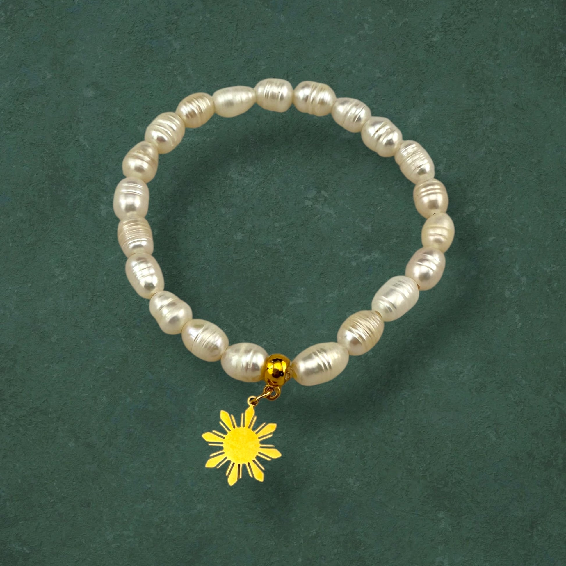 Aking Sinta Freshwater Pearl Stretch Bracelet with white beads and gold Araw charm displayed on dark green background, highlighting natural stone beads and sun motif