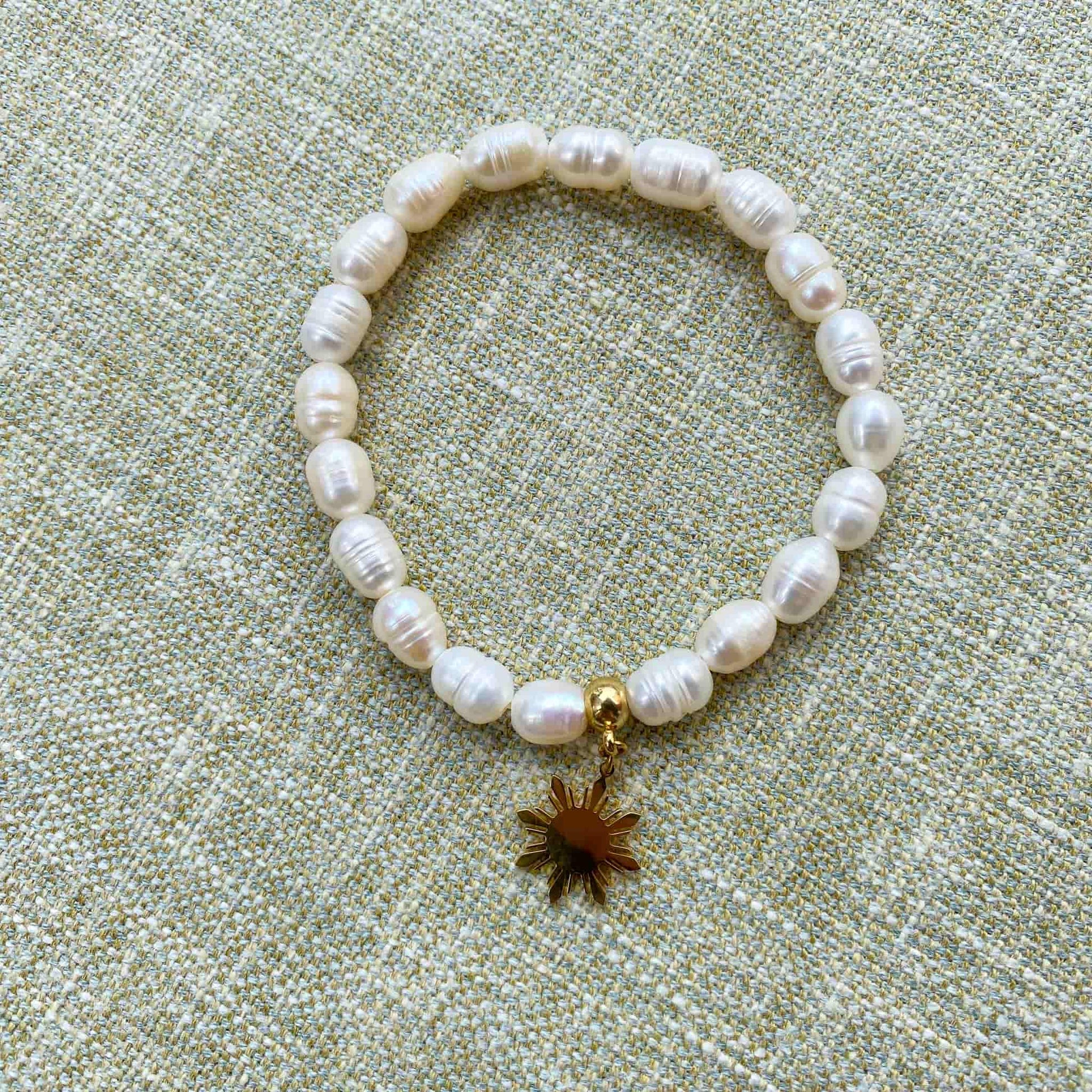 Aking Sinta Freshwater Pearl Stretch Bracelet with white beads and gold Araw charm displayed on gray woven textured background, highlighting natural stone beads and sun motif