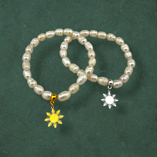 Aking Sinta Freshwater Pearl Stretch Bracelet with white beads, featuring gold and silver Araw sun charms, displayed on hand against dark green background