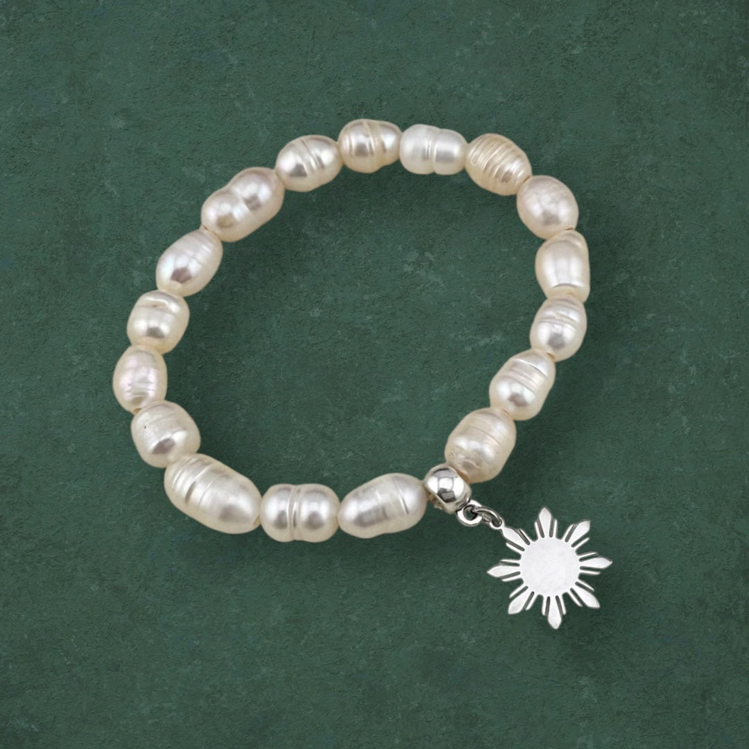 Aking Sinta Freshwater Pearl Stretch Bracelet with white beads and silver Araw charm displayed on dark green background, highlighting natural stone beads and sun motif