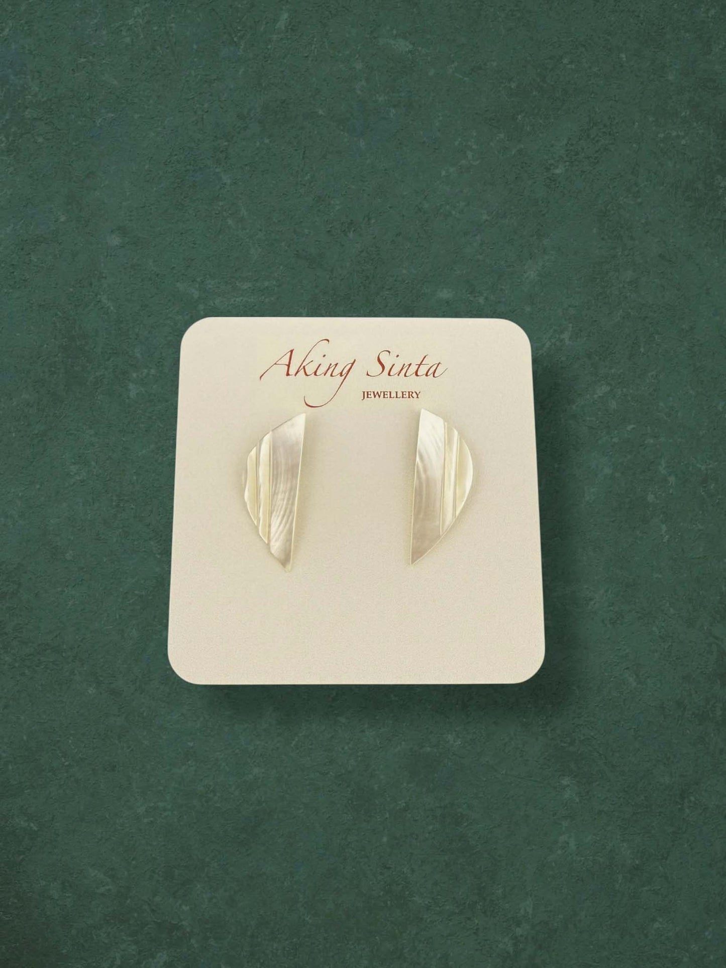A pair of Aking Sinta Mother of Pearl stud earrings in a sleek, geometric crescent shape. These minimalist iridescent shell earrings feature a vertical ribbed texture on a branded white card