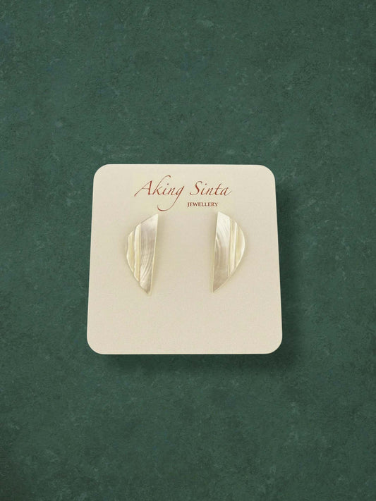 A pair of Aking Sinta Mother of Pearl stud earrings in a sleek, geometric crescent shape. These minimalist iridescent shell earrings feature a vertical ribbed texture on a branded white card